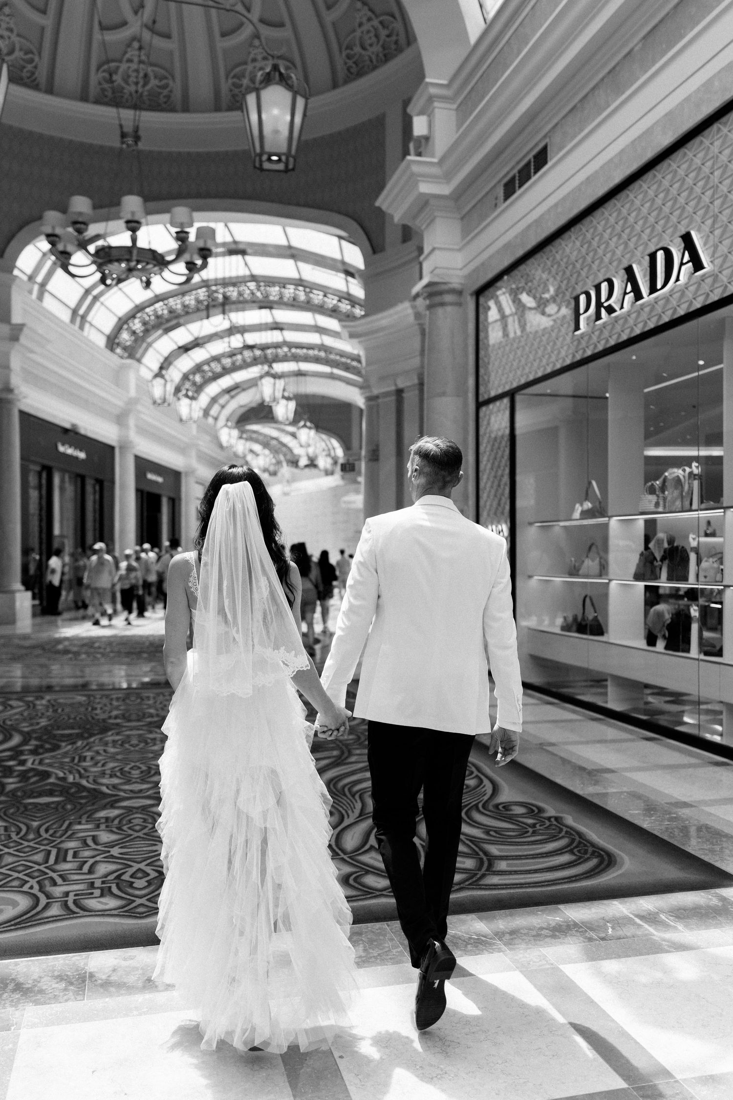 Bride and groom walking through an elegant shopping mall with ornate ceiling and luxury stores.