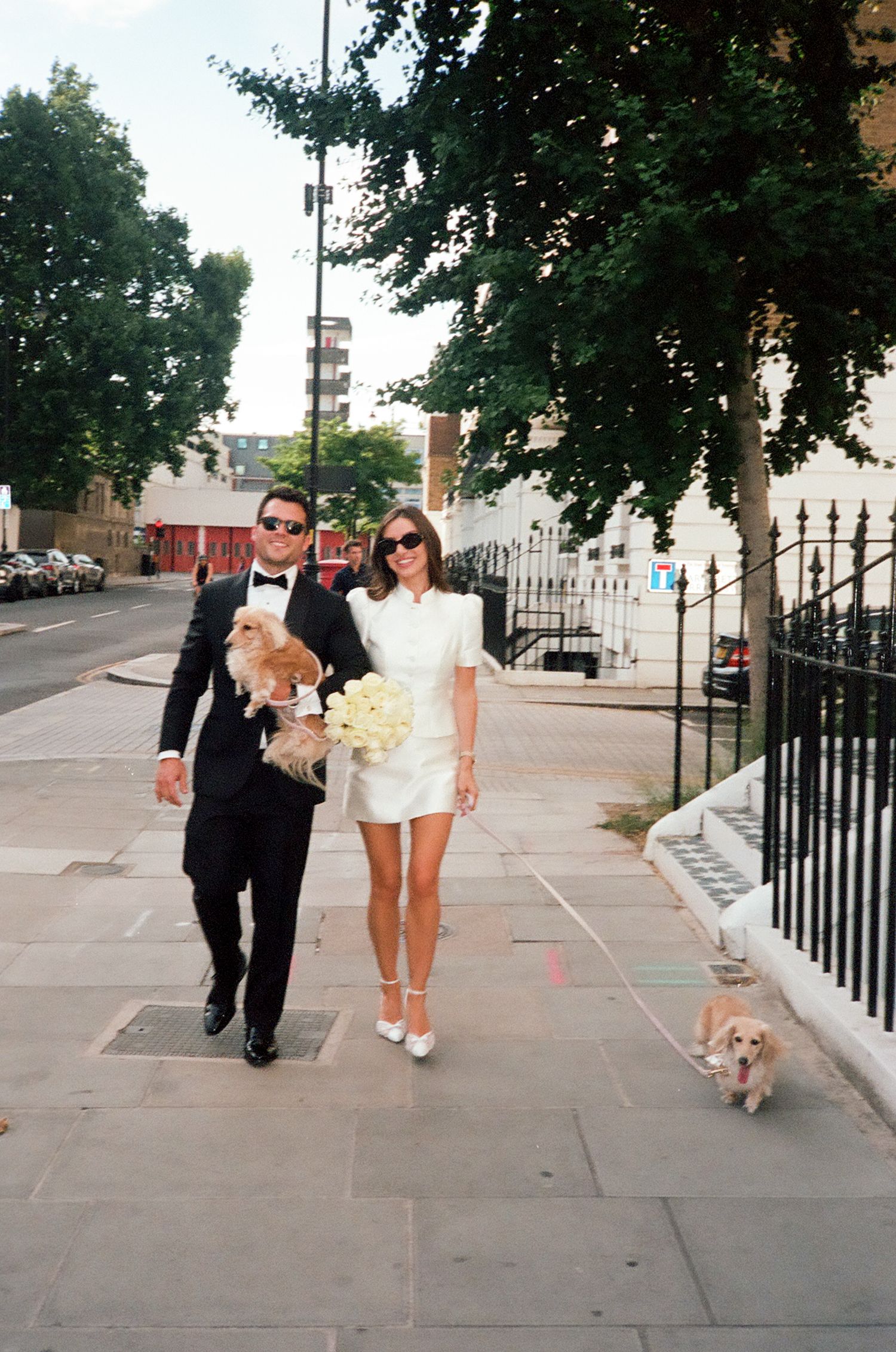 Bride in white mini dress and groom in black tuxedo walking two Corgi dogs on a sunny city sidewalk.