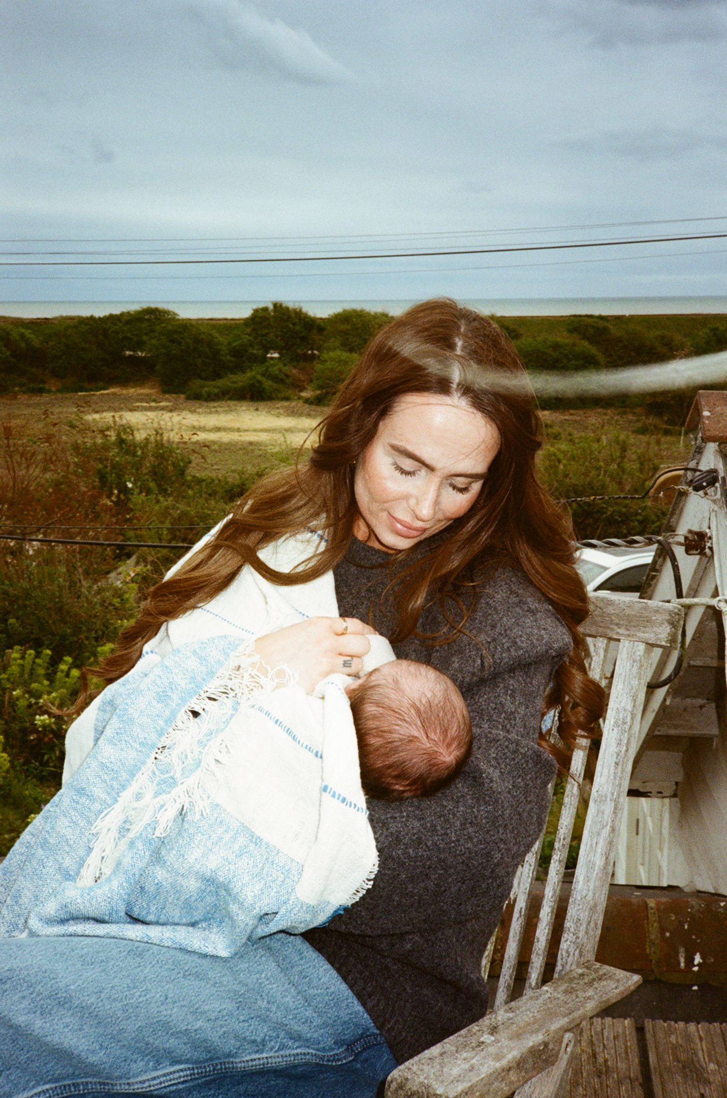 A mother tenderly breastfeeds her newborn baby outdoors on a wooden bench with a scenic coastal landscape behind her.
