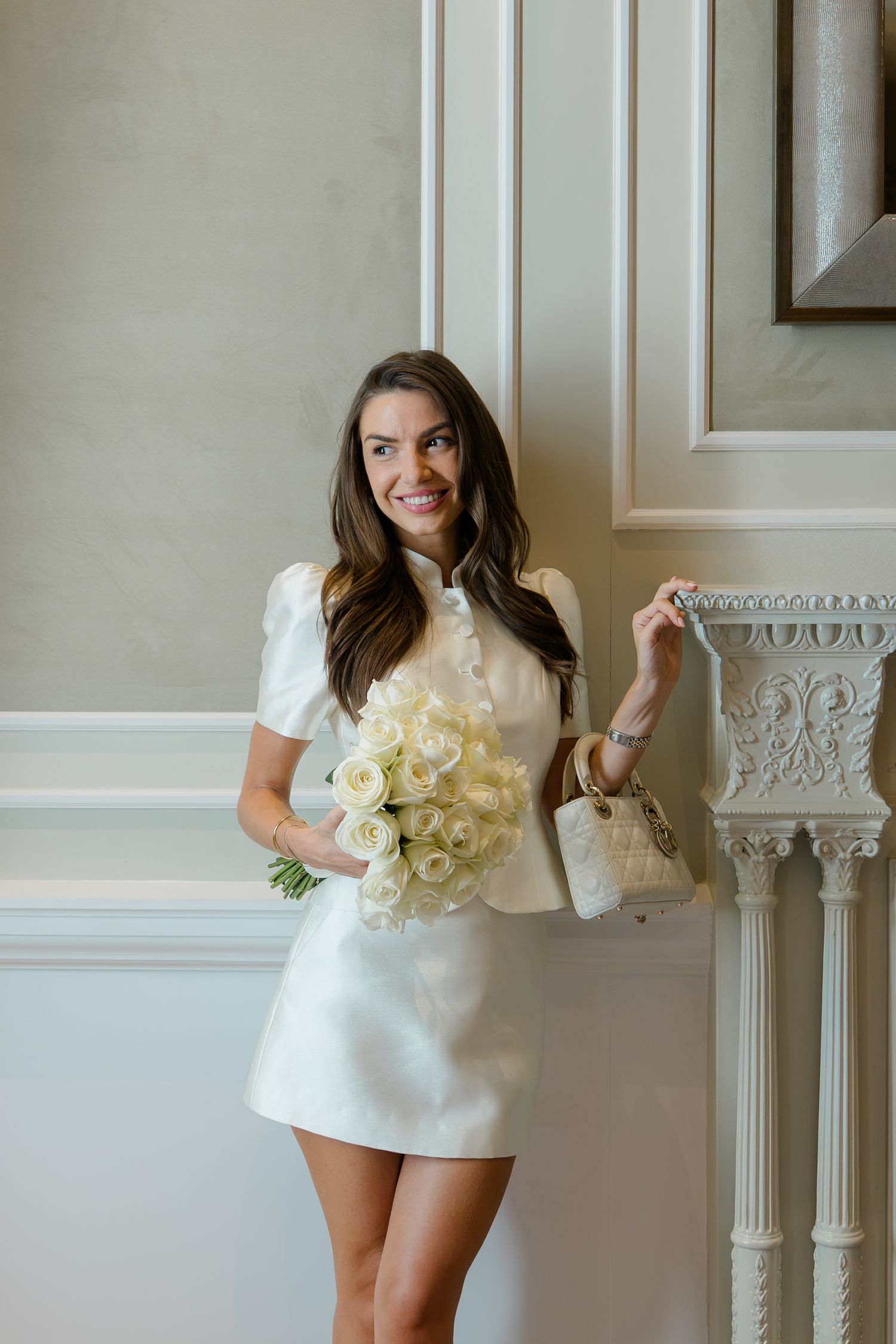 Elegant bride in white mini dress with puff sleeves holds white rose bouquet, posing by ornate white wall molding.