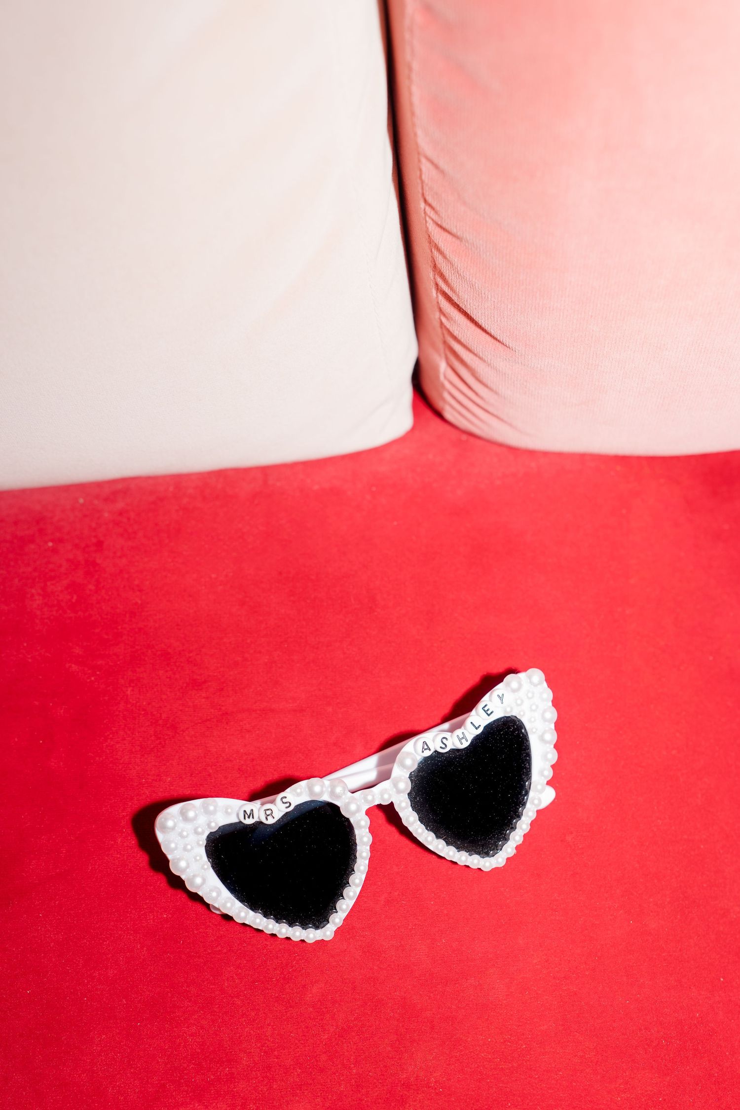 White heart-shaped sunglasses with pearl trim spelling MRS on a red velvet sofa cushion.