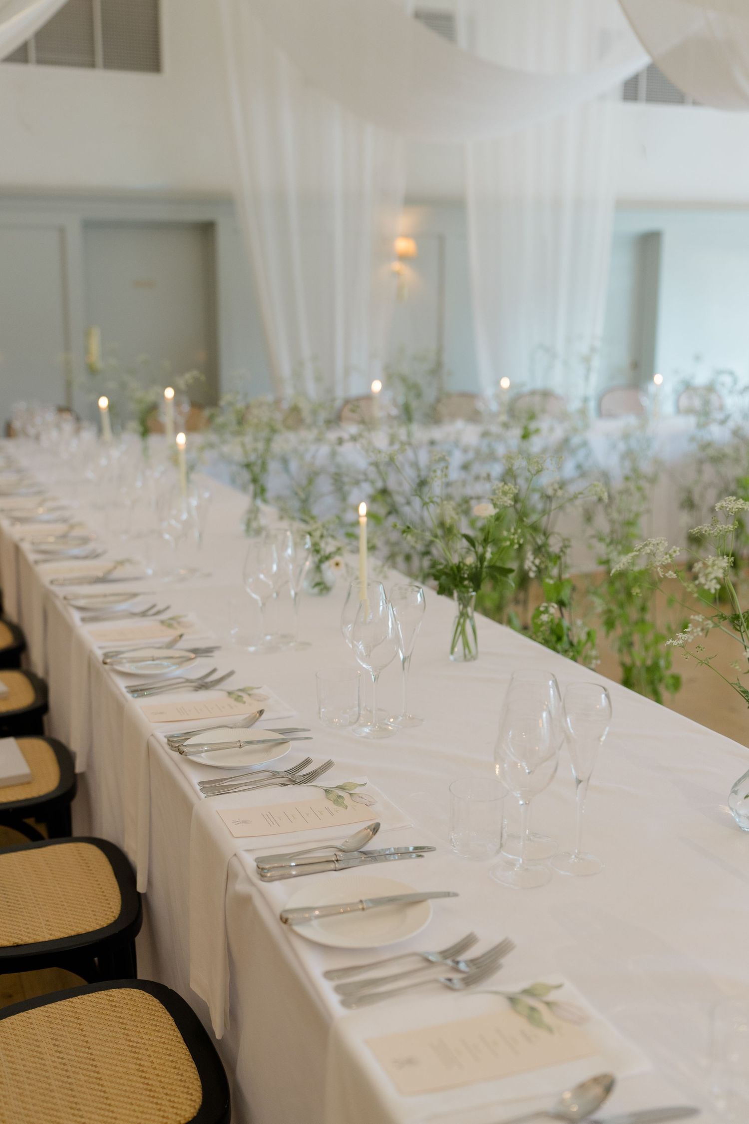 Elegant wedding reception long table set with white linens, crystal glasses, candles, and delicate greenery floral arrangements.