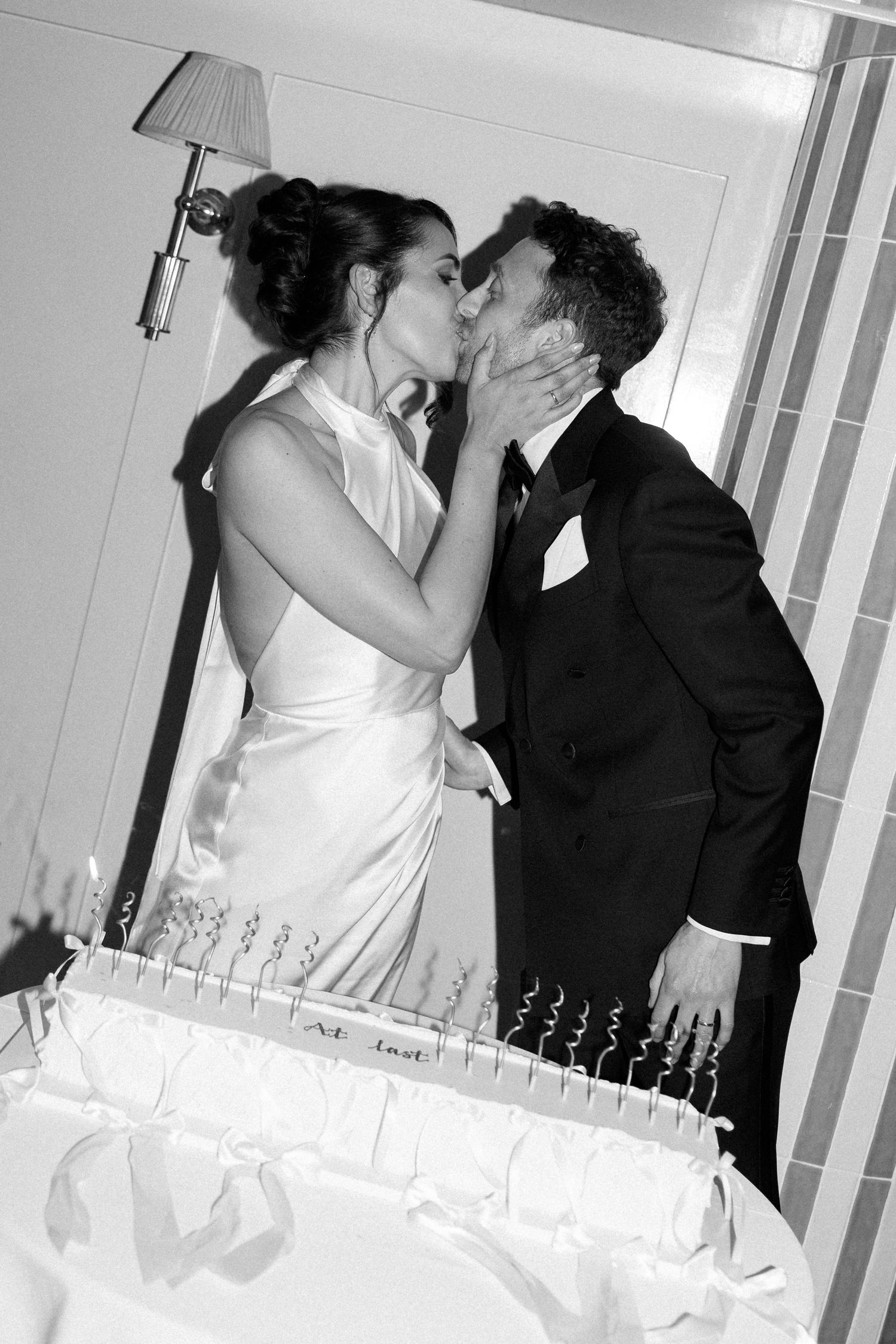 Black and white photo of bride and groom sharing a kiss over their wedding cake.