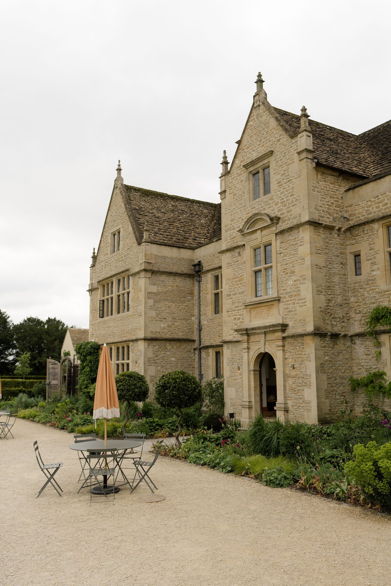 Historic Cotswold stone manor house with outdoor cafe seating, manicured gardens, and Gothic architectural details on overcast day.