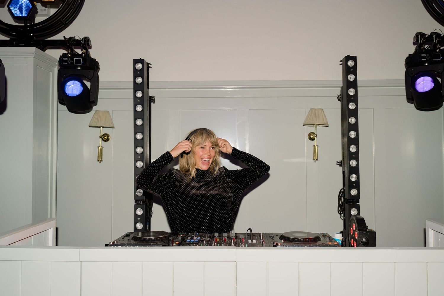 Female DJ performing at wedding reception behind decks with stage lighting in white paneled room.