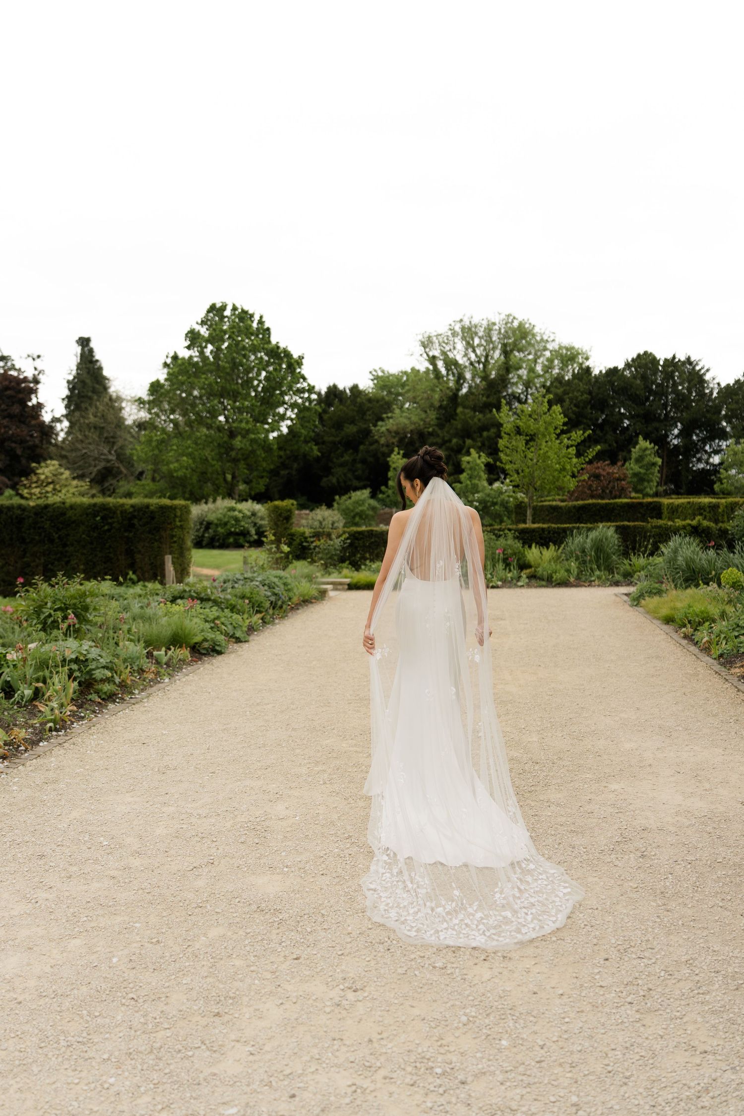 Bride in lace-trimmed cathedral veil walks through formal garden pathway lined with manicured hedges.