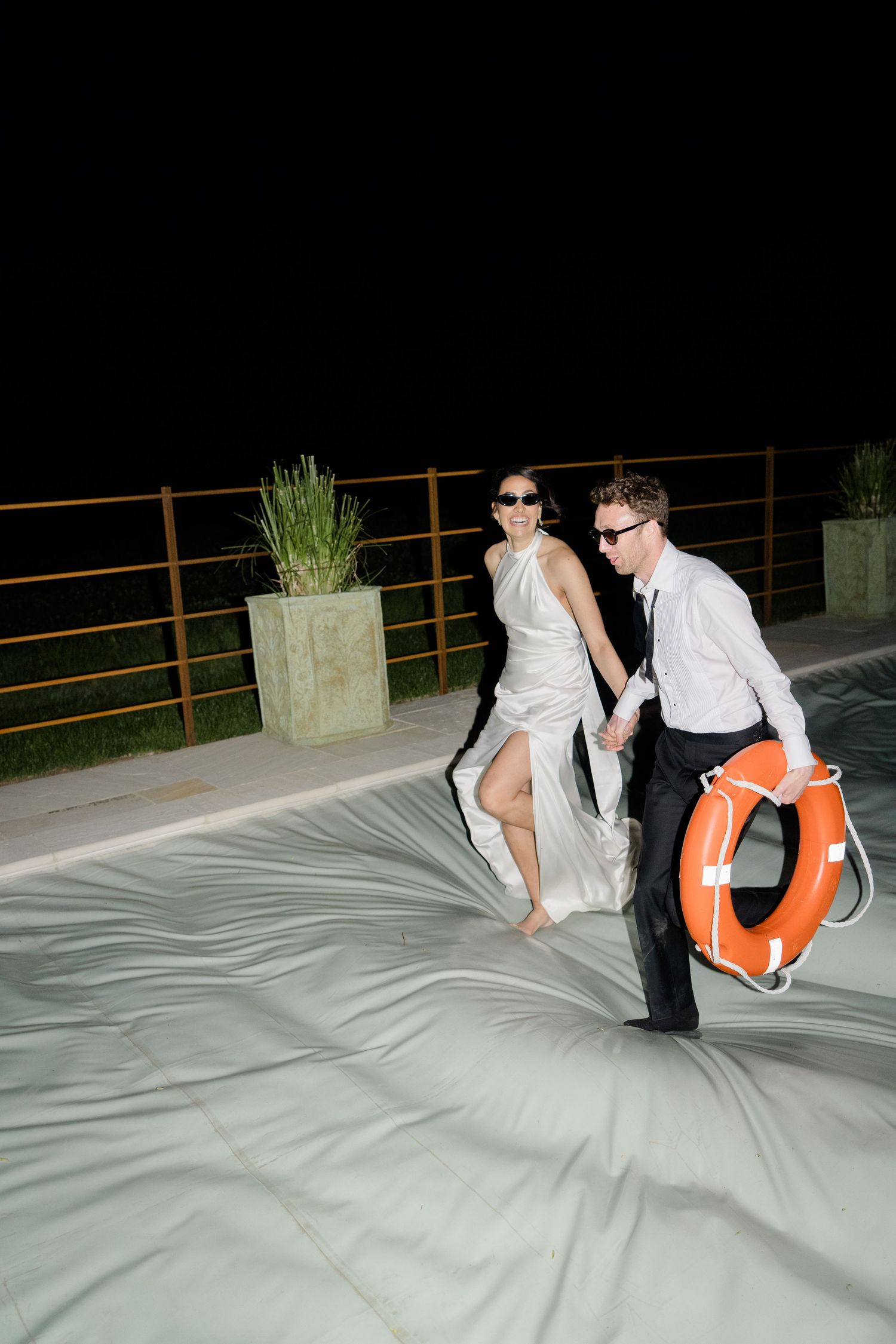 Newlyweds in sunglasses laugh while running across pool cover at night holding orange life preserver.