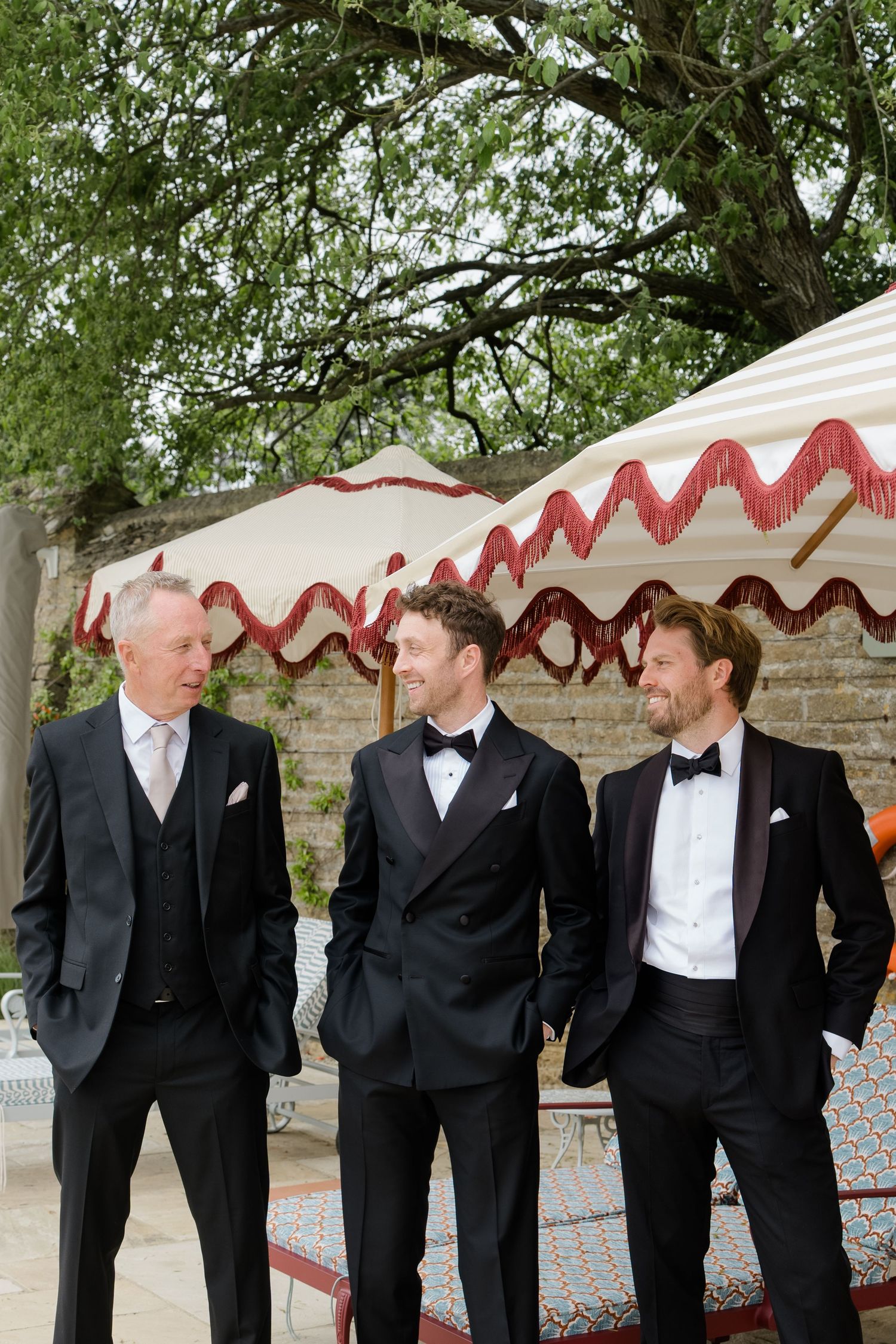 Three men in black tuxedos with bow ties stand outdoors at a wedding venue beneath a striped canopy.