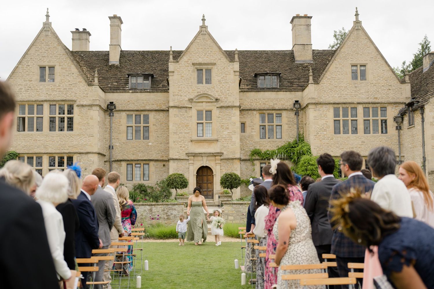 Outdoor wedding ceremony on manicured lawn before grand Cotswolds stone manor house with seated guests.