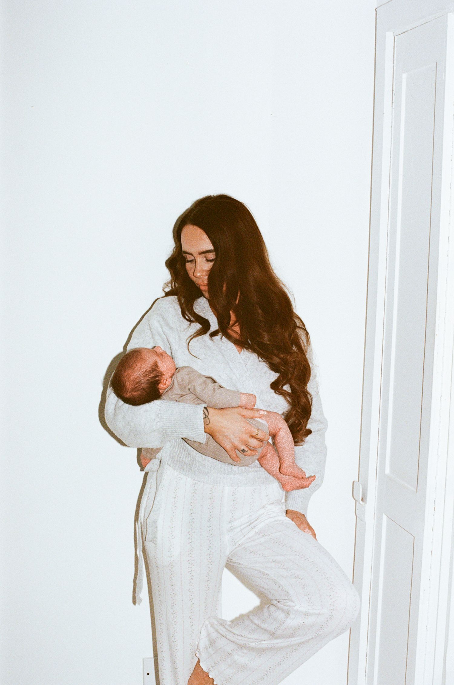 A woman in white loungewear cradles a newborn baby against a bright white background.