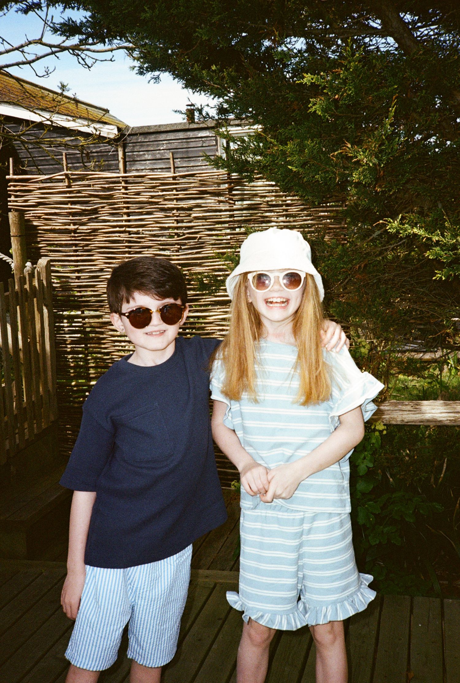 Two kids in sunglasses pose outdoors, boy in navy tee and striped shorts, girl in striped ruffle romper and bucket hat.