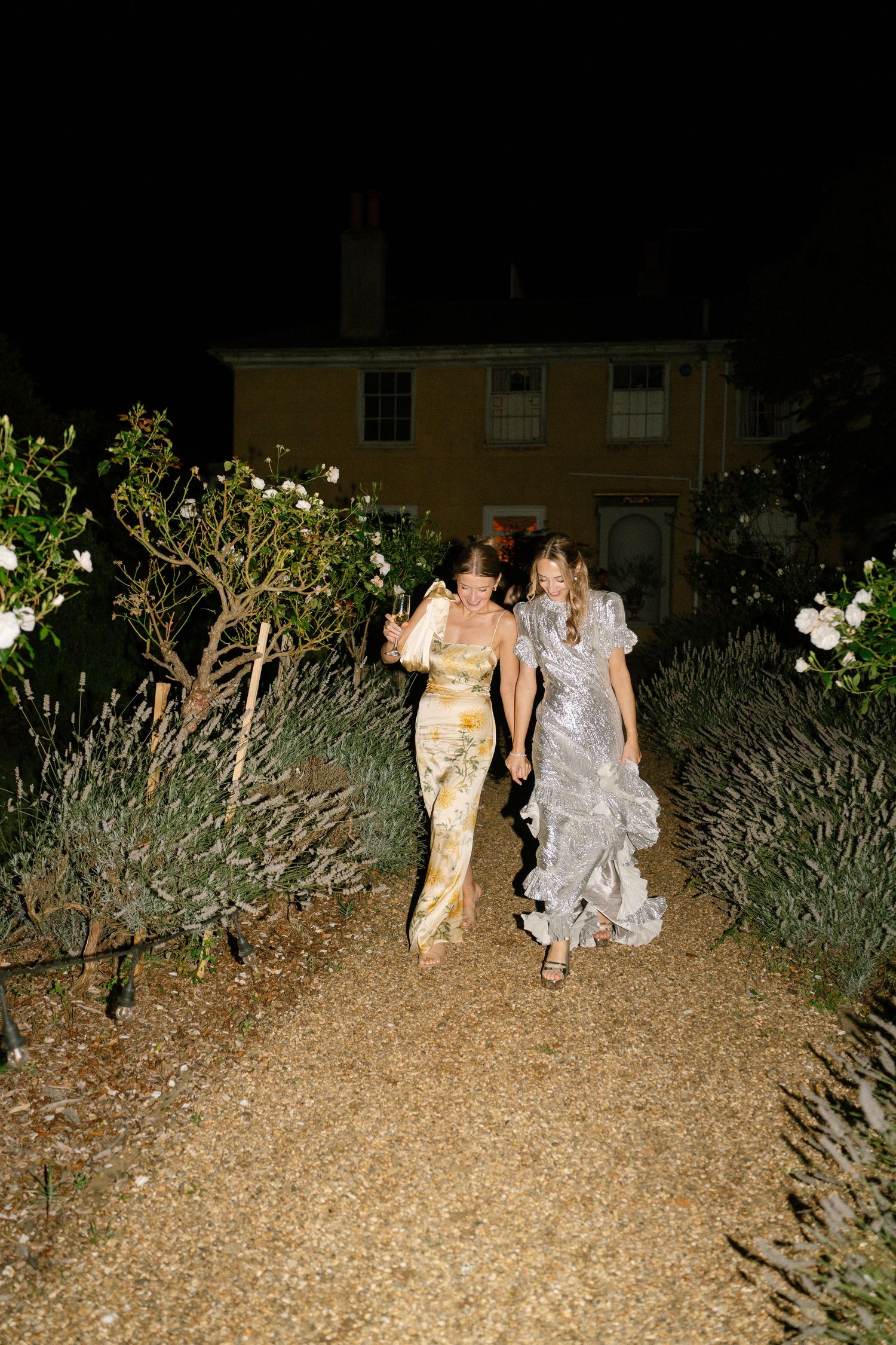 Wedding guests walk through a garden path at night in formal attire.