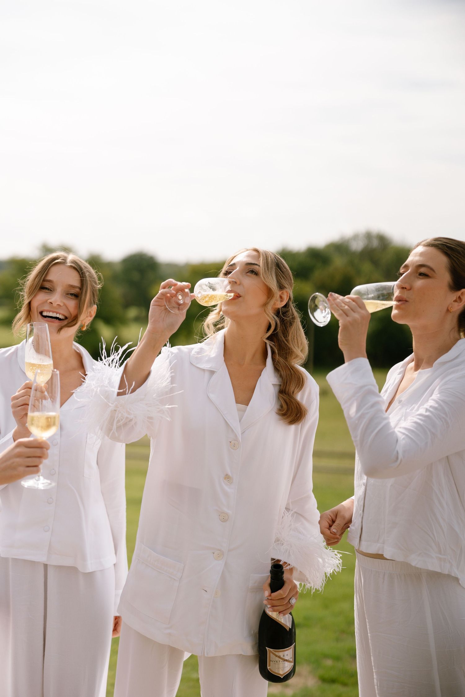 Friends in white outfits share champagne and laughter during an outdoor celebration.