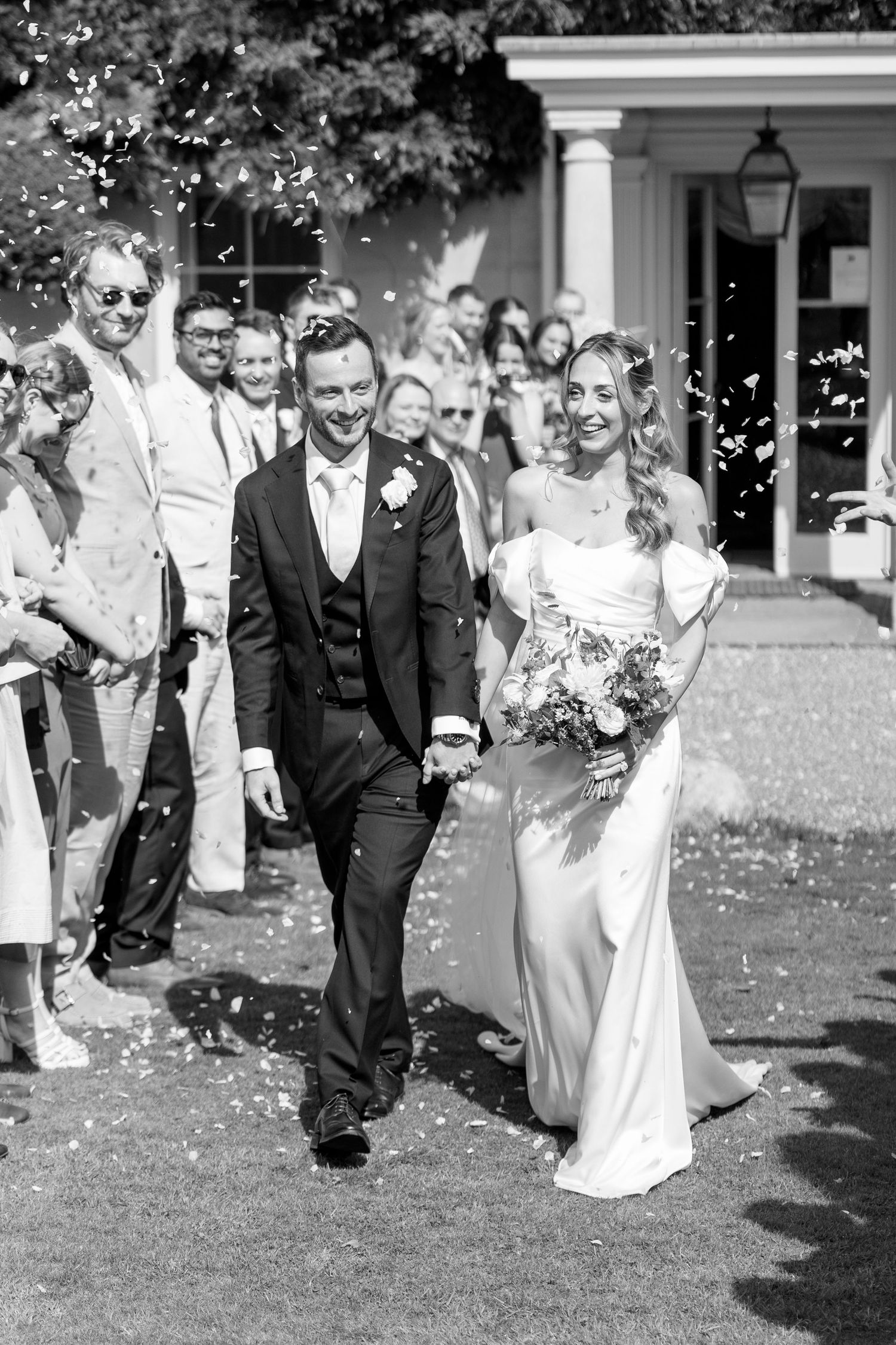 Newlyweds walk through shower of confetti as guests celebrate at outdoor wedding venue.
