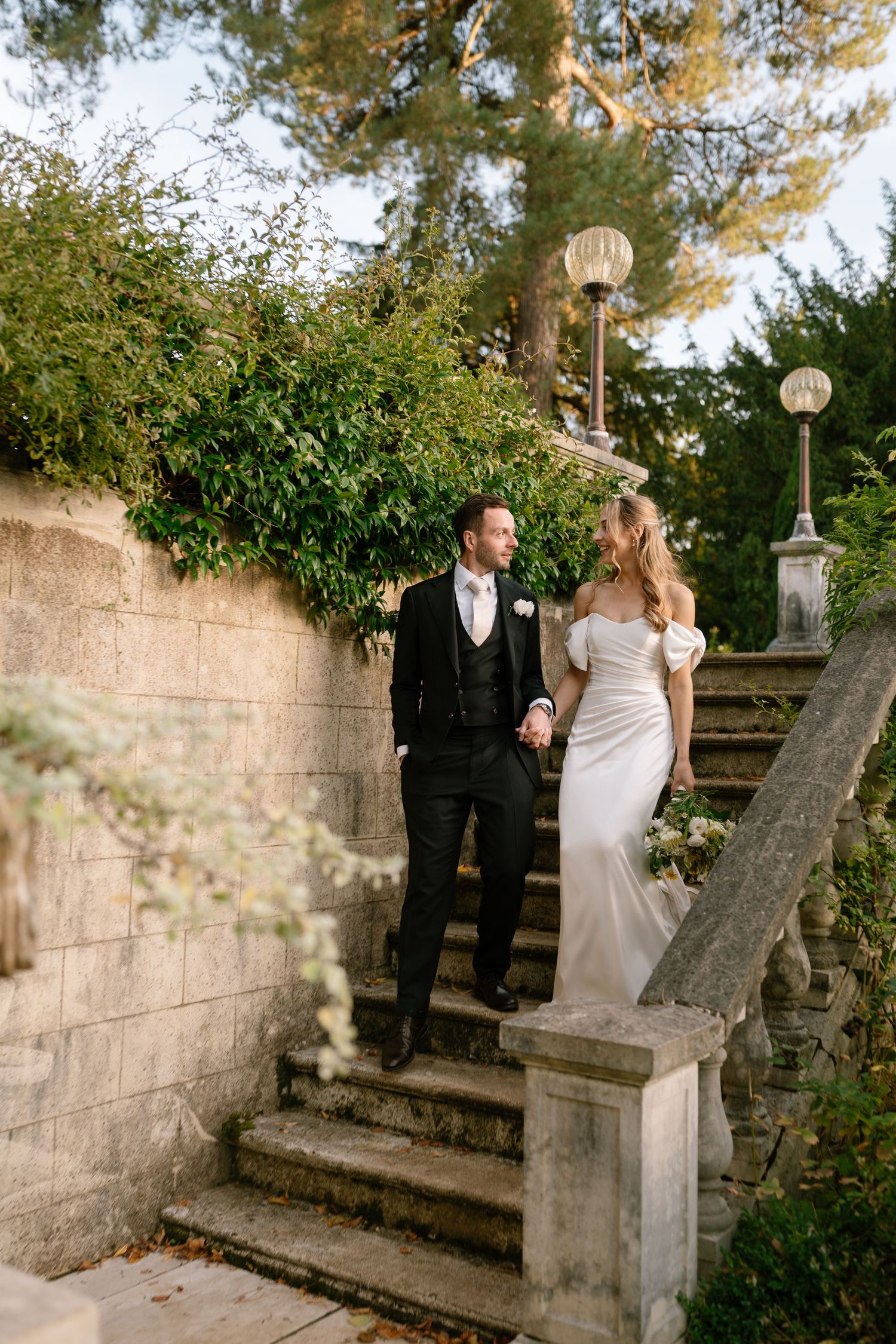 A wedding couple stands on elegant stone stairs surrounded by lush greenery and decorative lampposts.