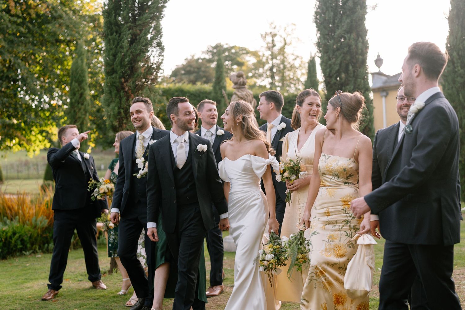 Wedding party members socialize outdoors during sunset with cypress trees framing the scene.