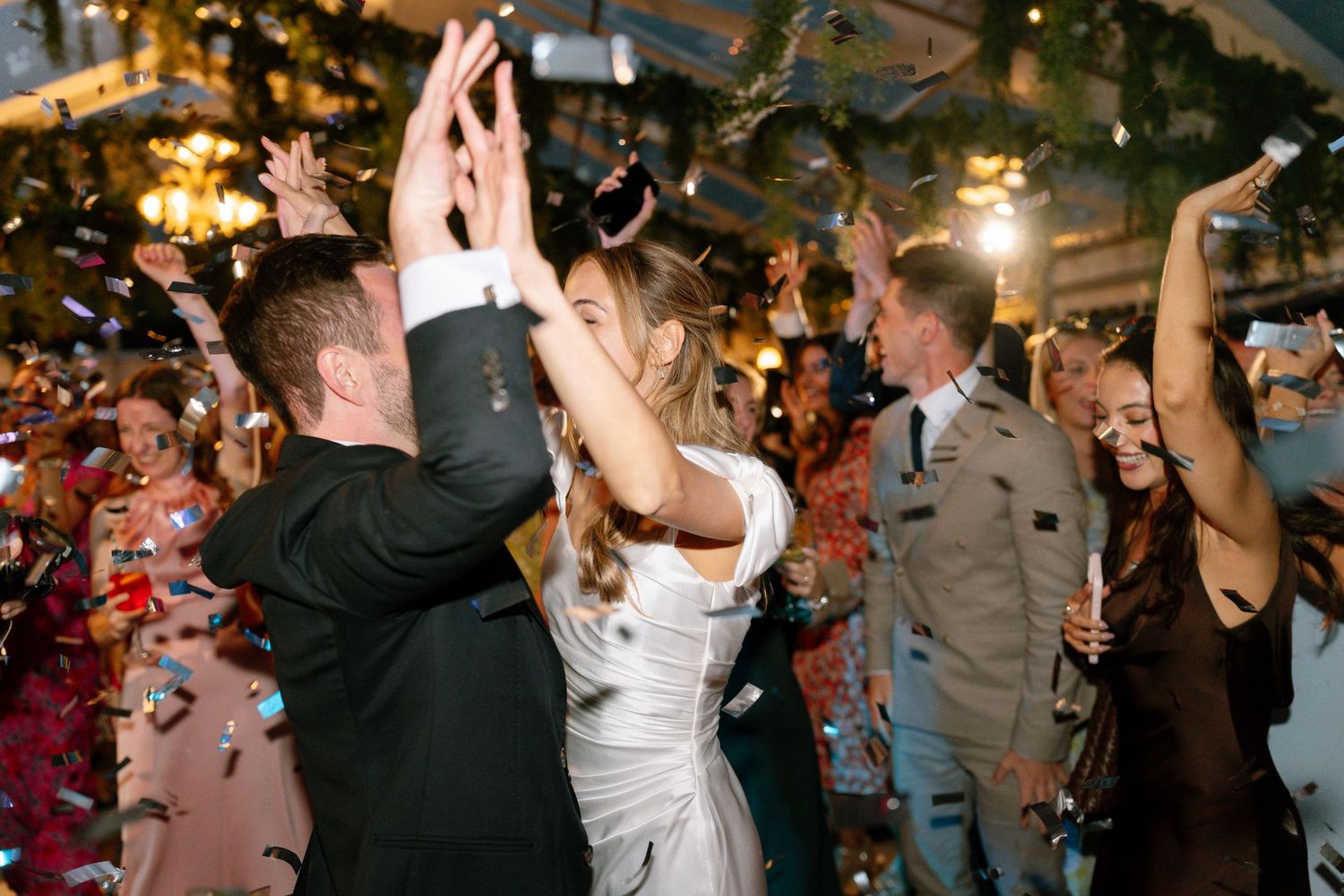 Wedding celebration with guests dancing and raising hands under twinkling lights.