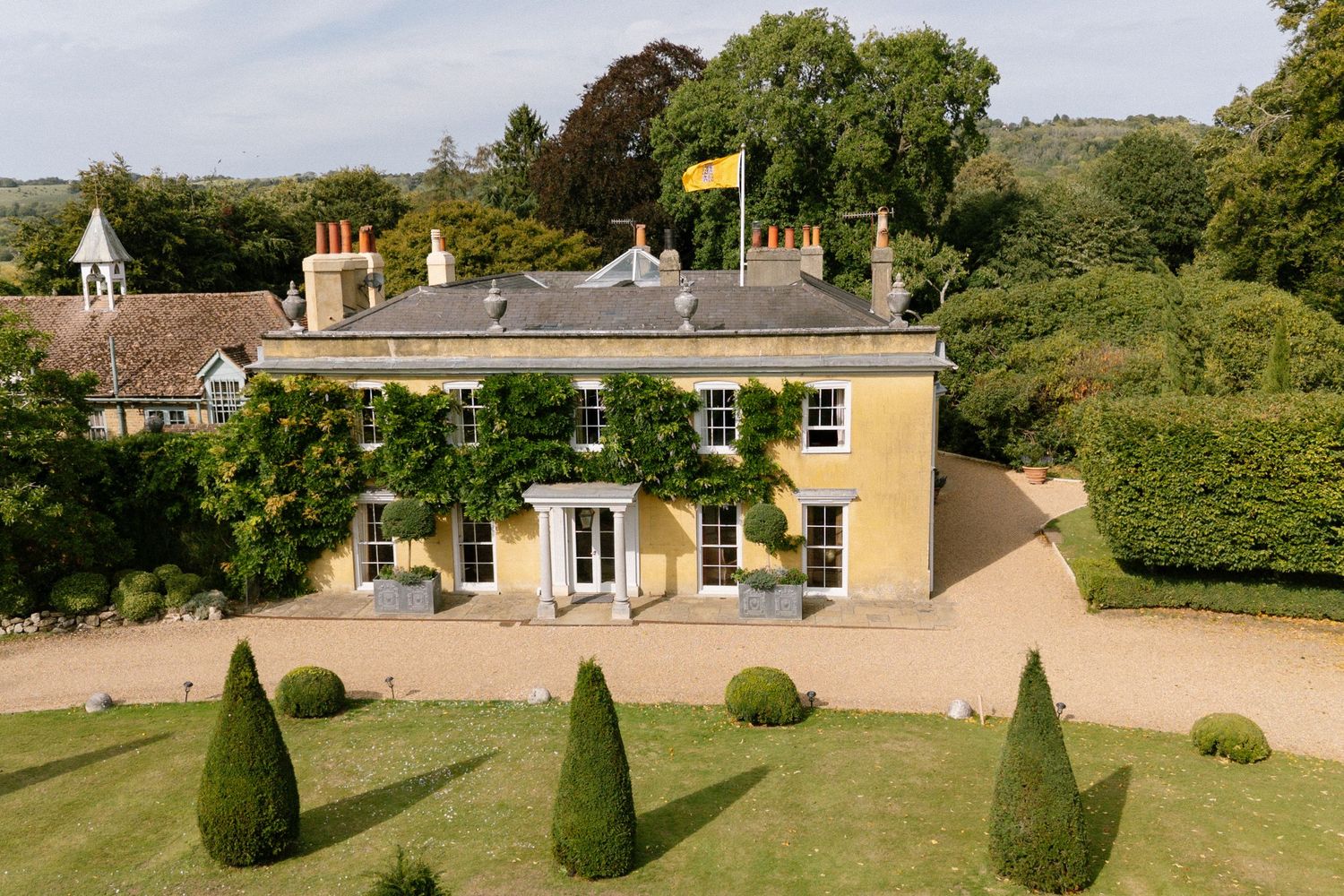 Elegant yellow country manor house with manicured gardens and gravel driveway in rural setting.