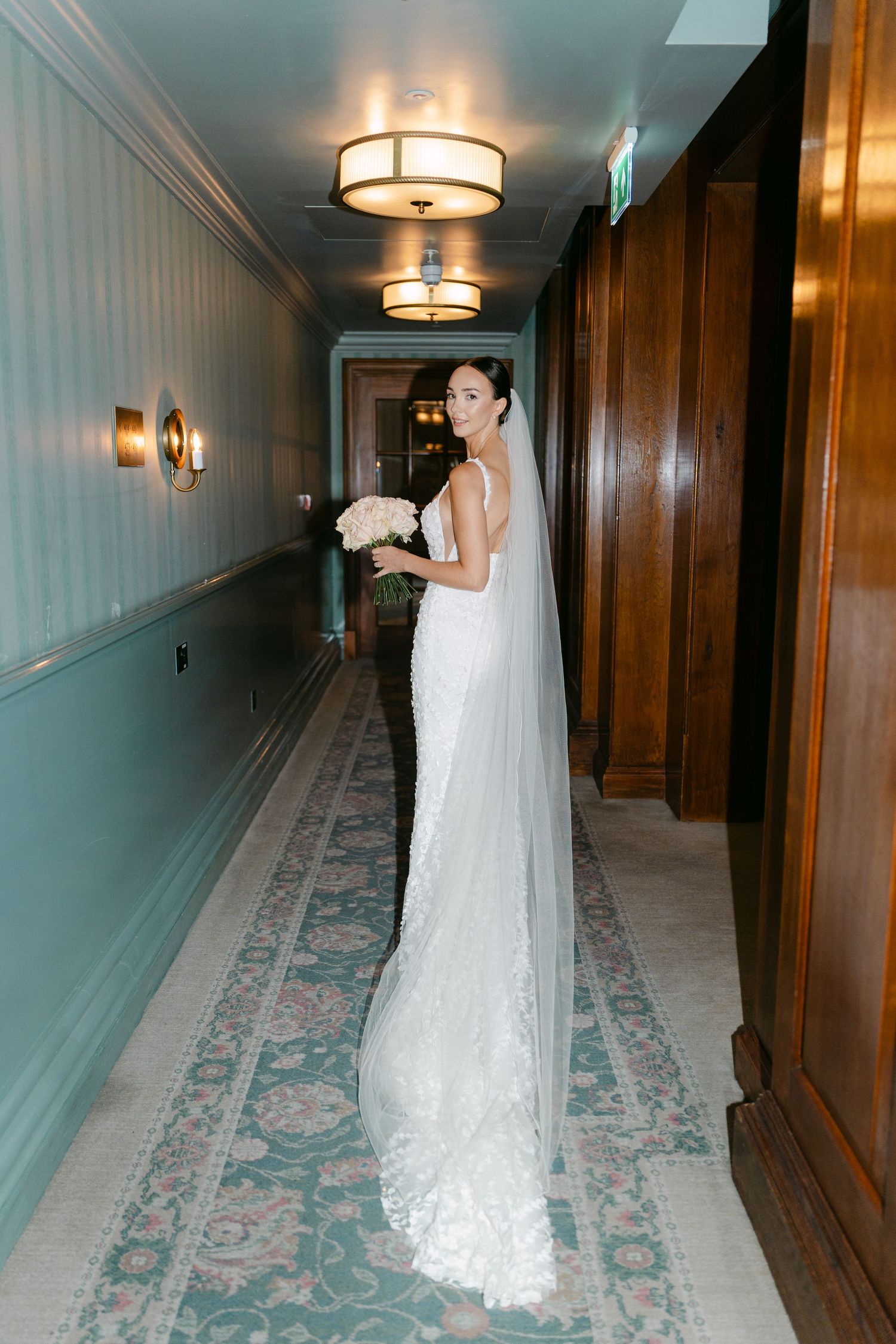 A bride in flowing white dress walks and poses along a carpeted hotel hallway with wood paneling.