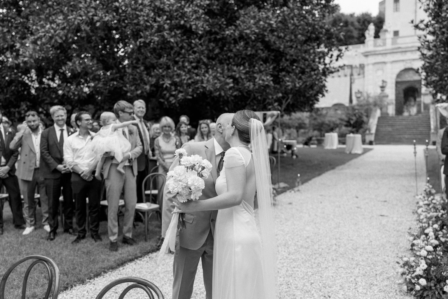 Wedding ceremony guests gather on a gravel path leading to a beautiful estate building.