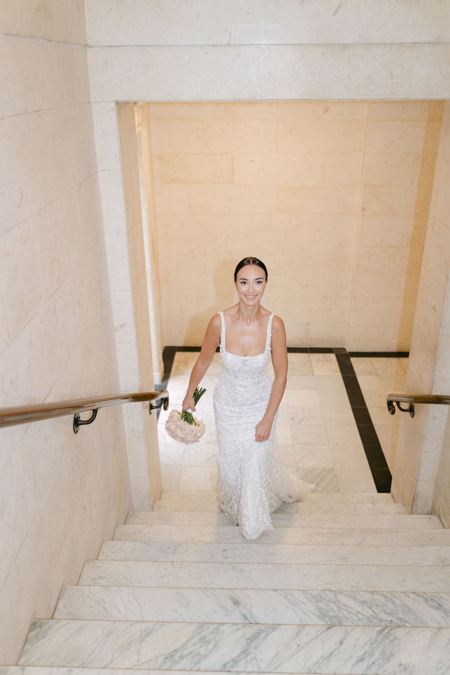 A bride in white lace gown ascends marble staircase holding bouquet.