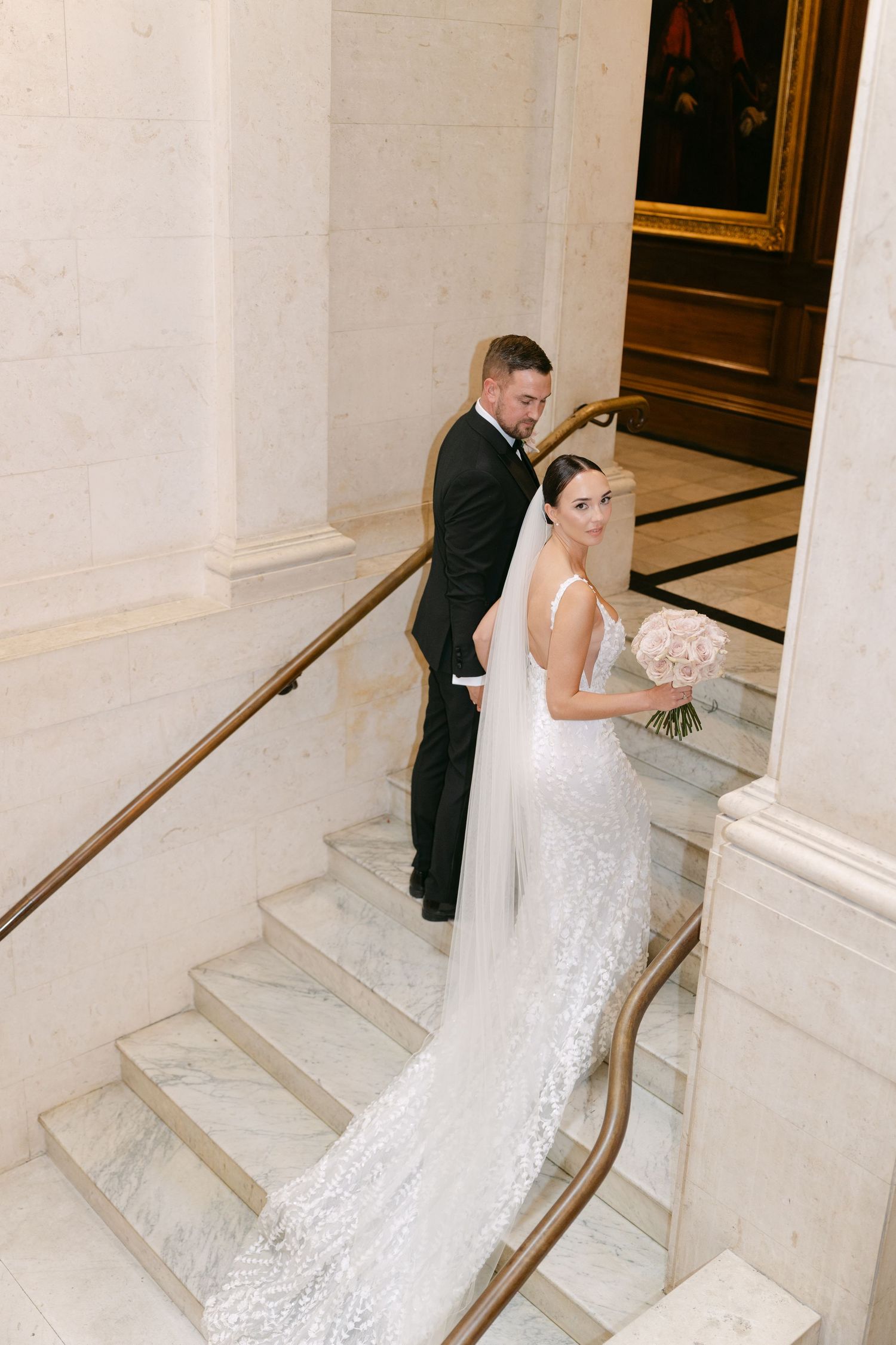 Wedding couple stands on marble steps with bride's detailed lace train flowing behind.