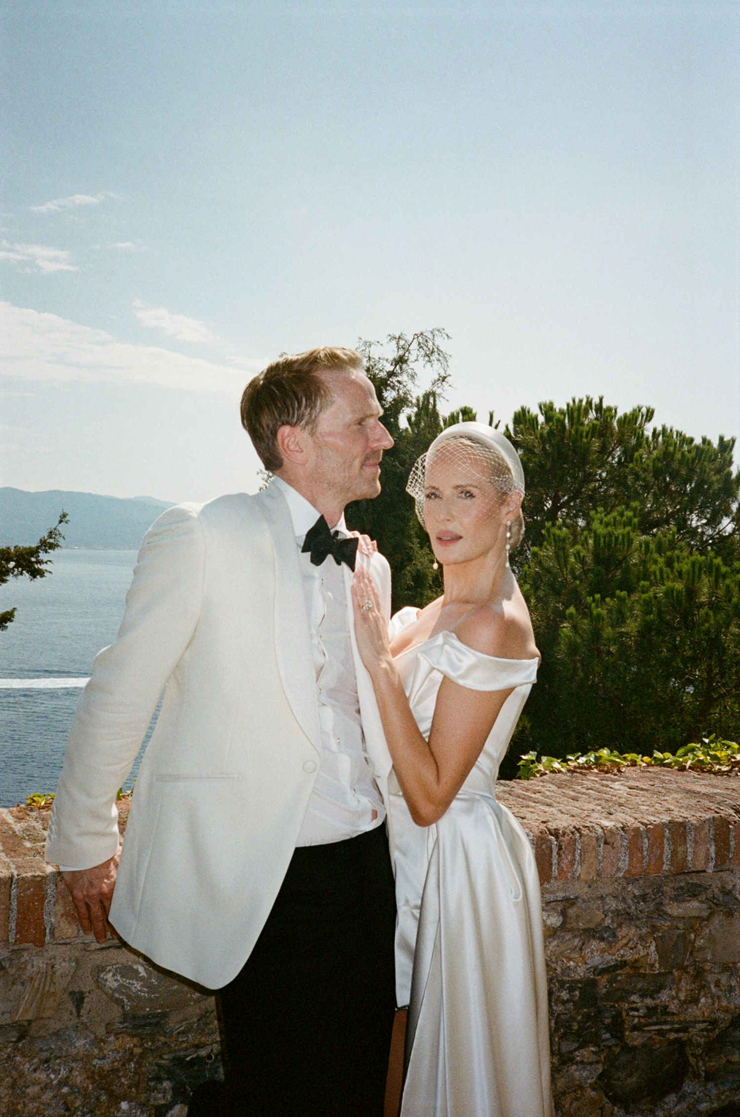 Elegant bride and groom in white tuxedo posing by stone wall with ocean view on sunny wedding day.