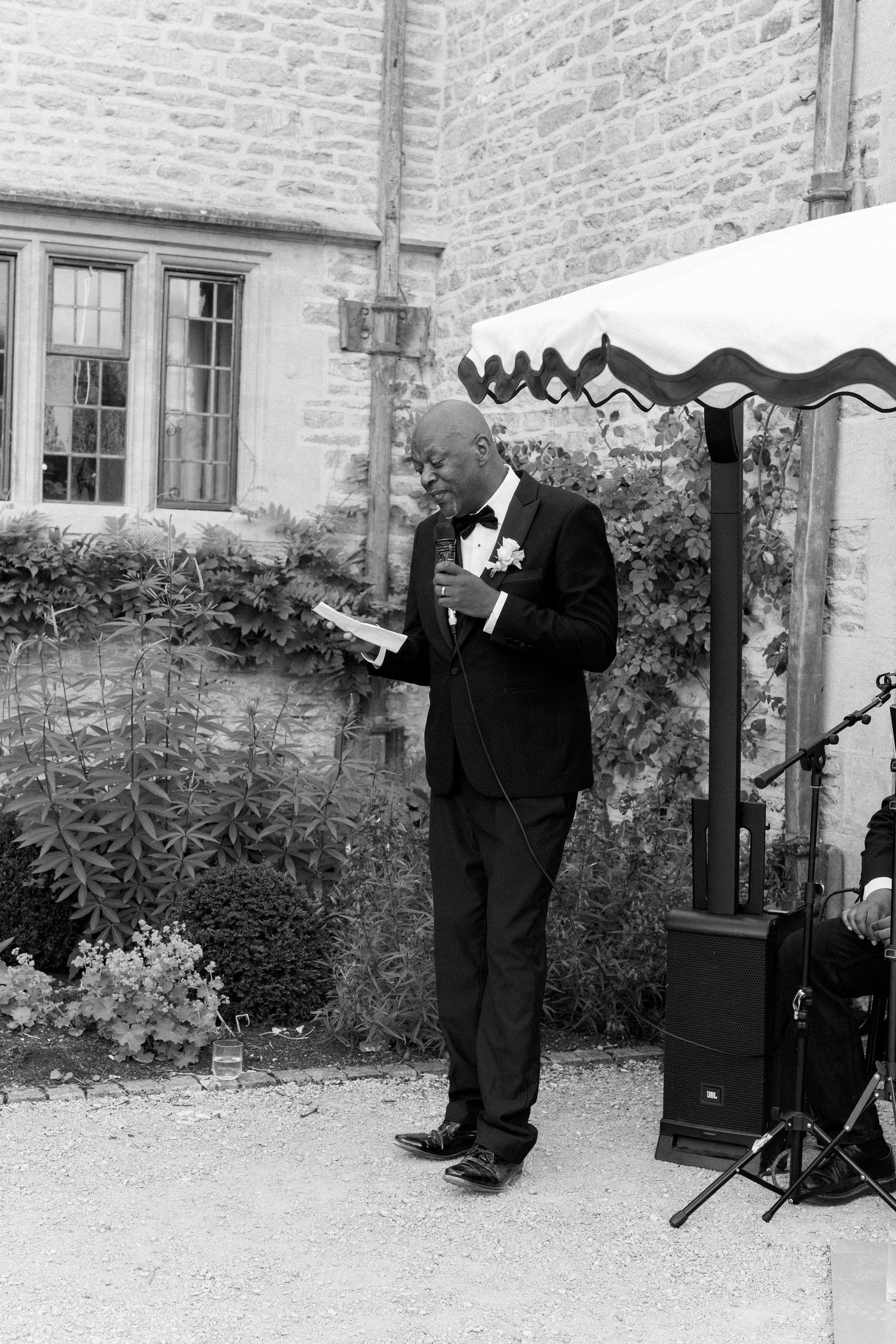 Elderly man in tuxedo delivers heartfelt speech at outdoor wedding near stone building and live band.