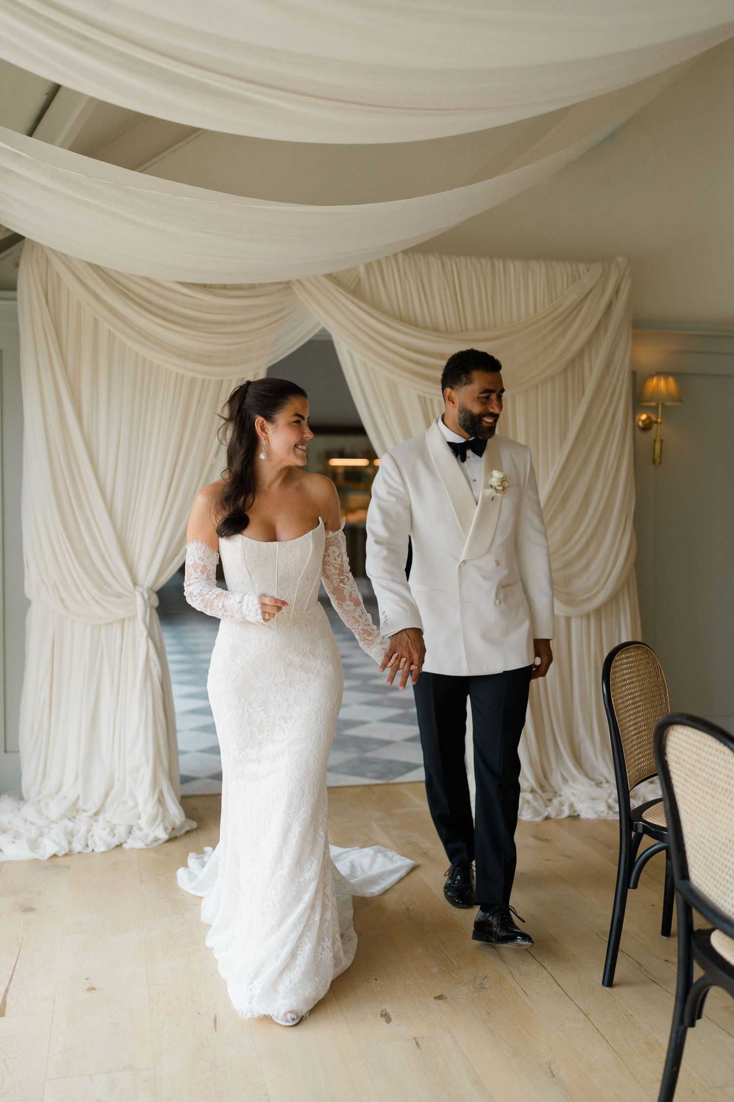 Bride in lace off-shoulder gown and groom in white tuxedo jacket walk hand in hand through draped arch.