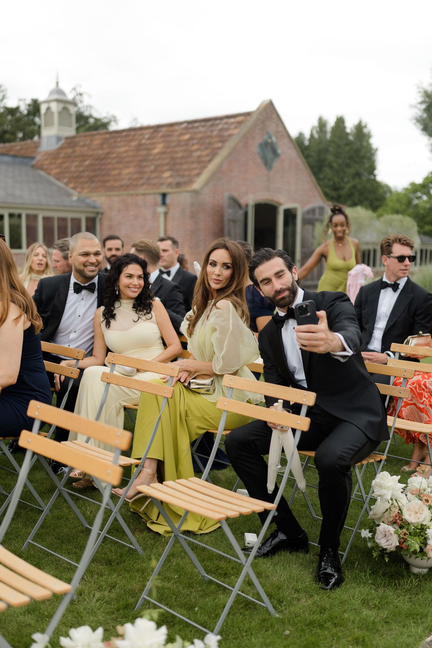 Wedding guests in formal attire take selfie while seated in outdoor ceremony chairs on lawn.