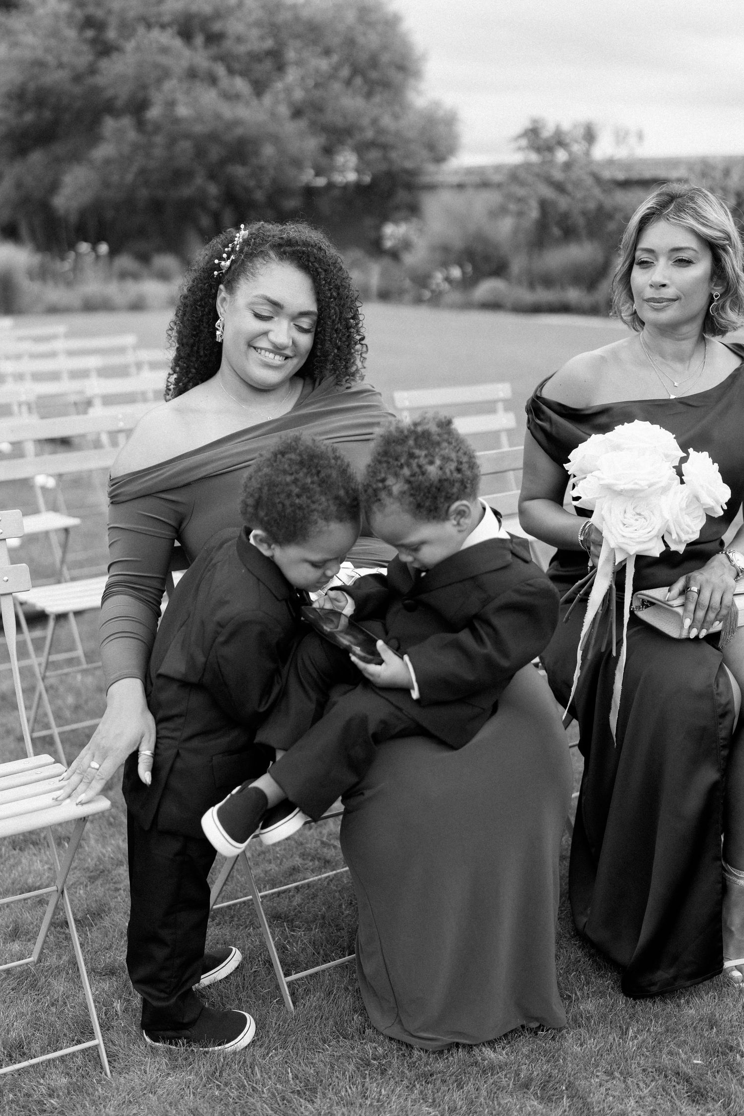 Two young boys in suits playfully interacting with bridesmaids holding white bouquet at outdoor wedding.