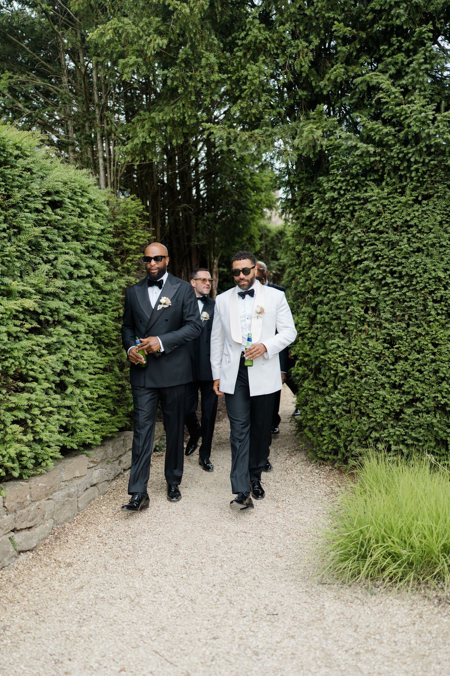 Groom in white tuxedo leads groomsmen in black suits along hedged garden pathway at wedding.
