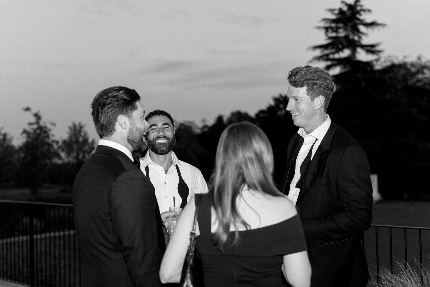 Black and white dusk photo of four wedding guests laughing and socializing outdoors near trees.
