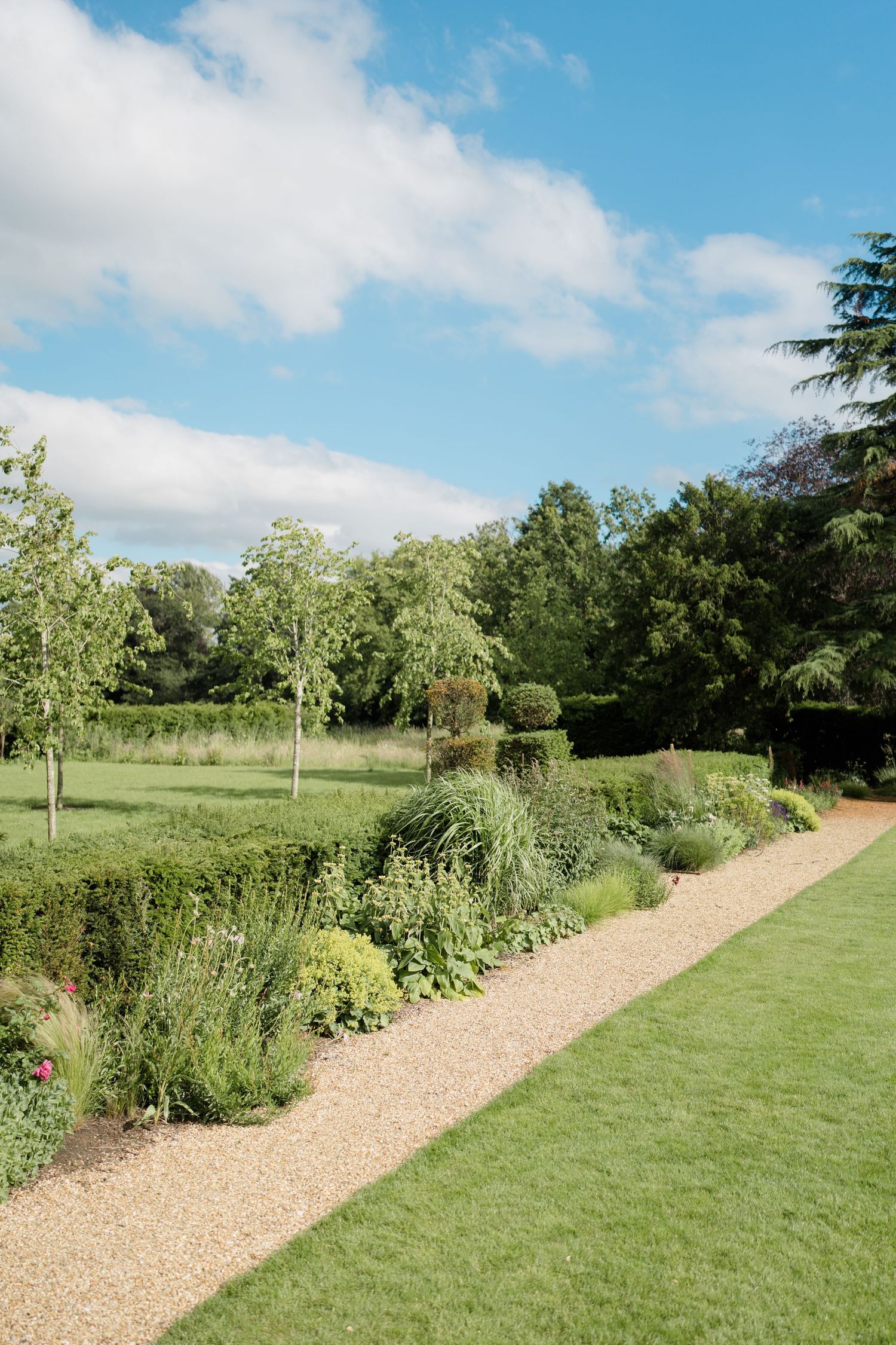 Manicured English garden with gravel path, flowering borders, and tall trees under blue sky.