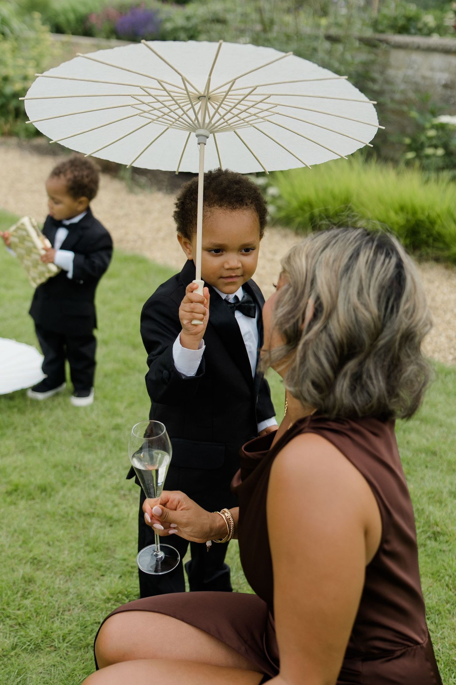 Young boy in tuxedo holding white parasol umbrella with wedding guest at garden reception.