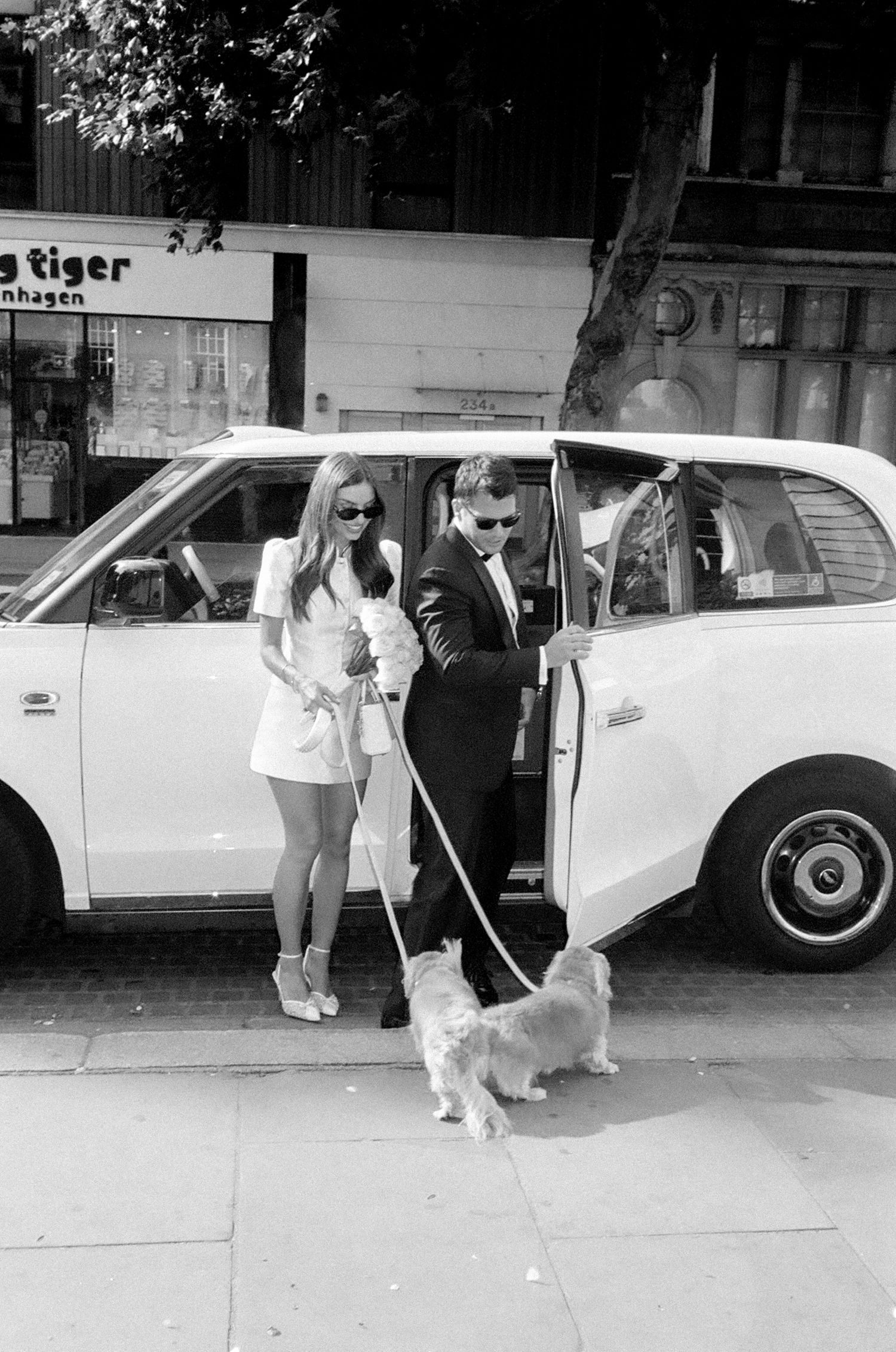 Stylish couple in black and white photo exiting a white car with a small white dog on a city street.