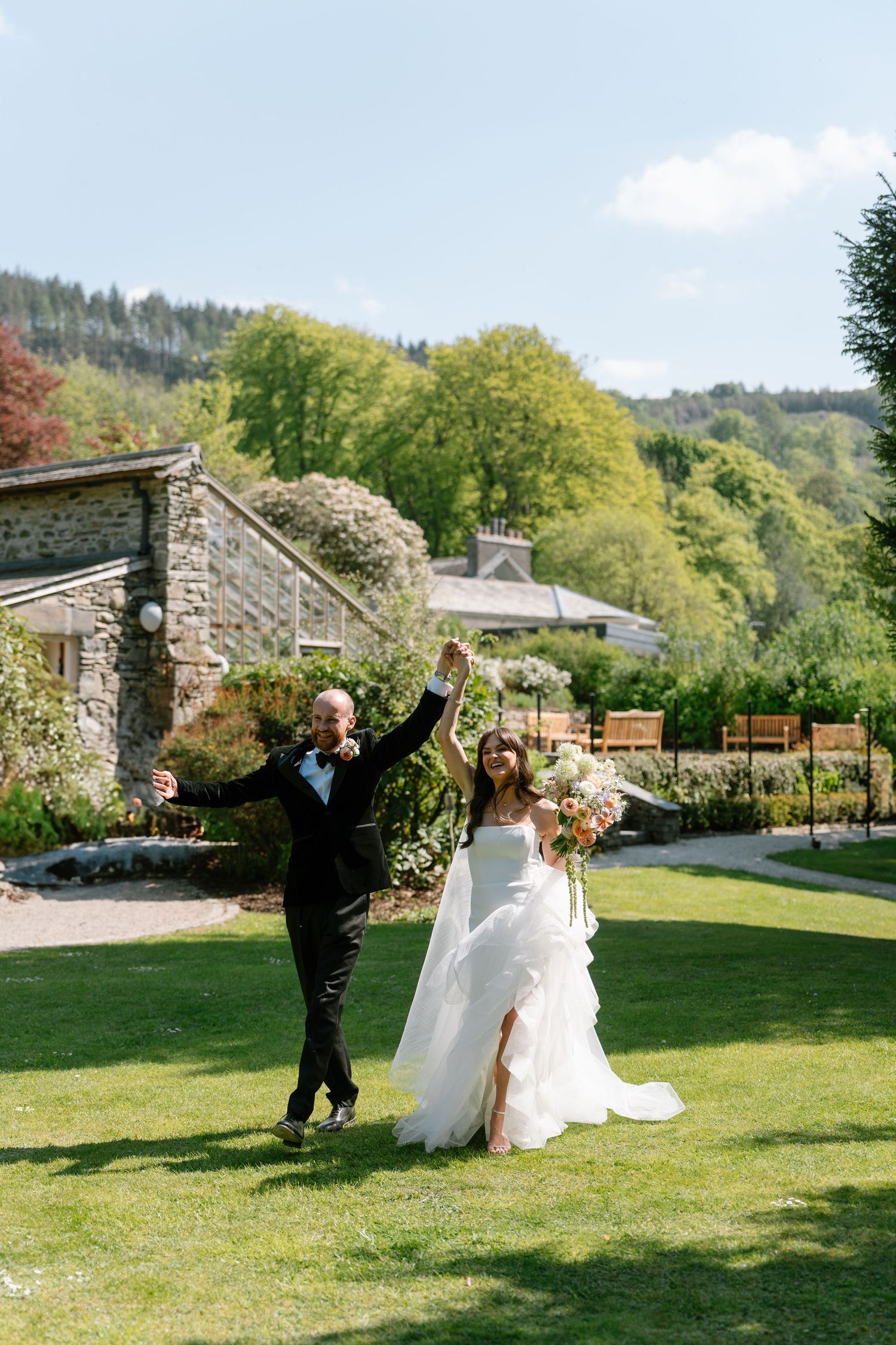 Newlyweds celebrate with raised arms on a beautiful garden lawn with stone buildings in background.