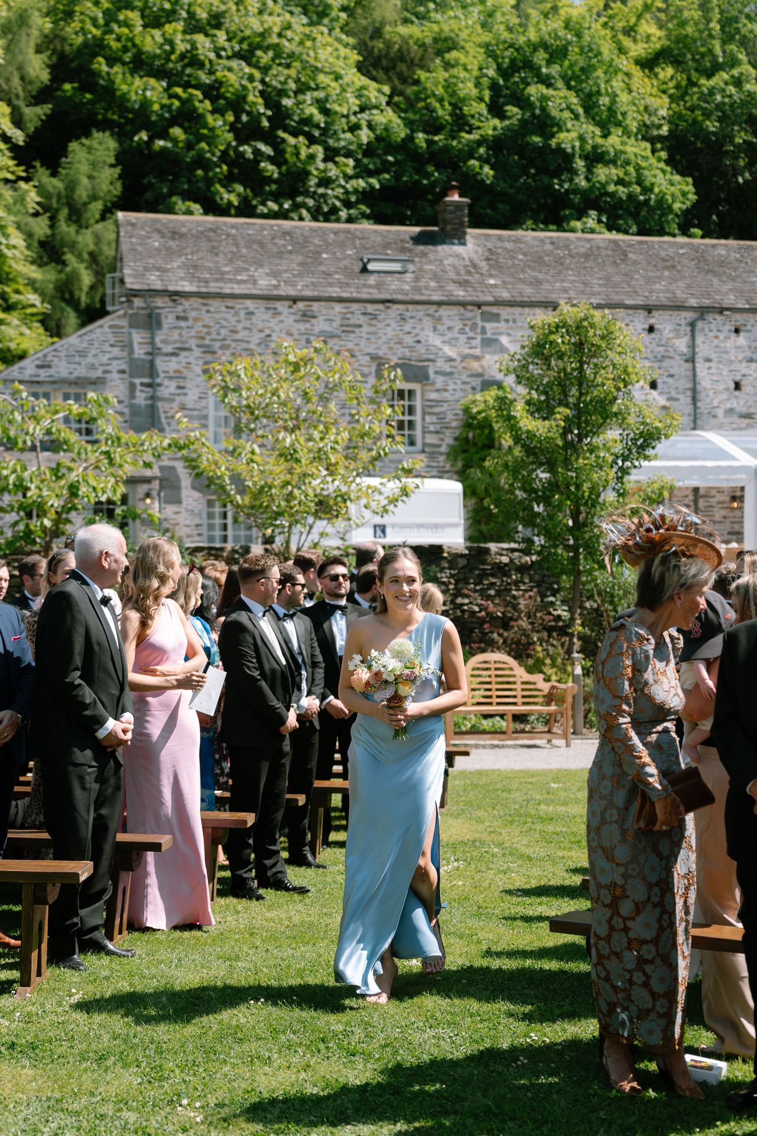 Wedding guests gather on a sunny lawn beside stone walls during an outdoor reception.