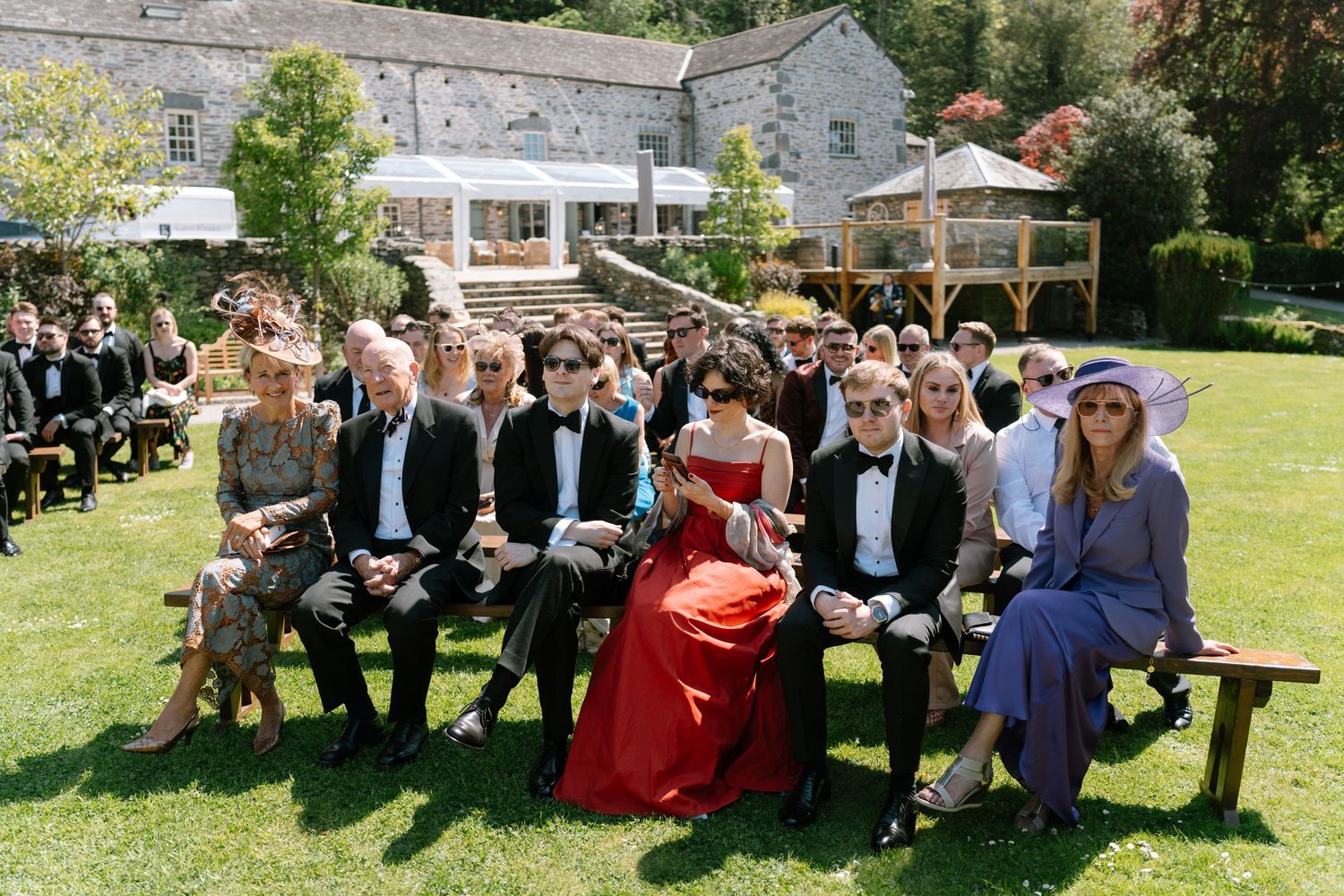 Wedding guests gather on benches and lawn during an outdoor celebration.