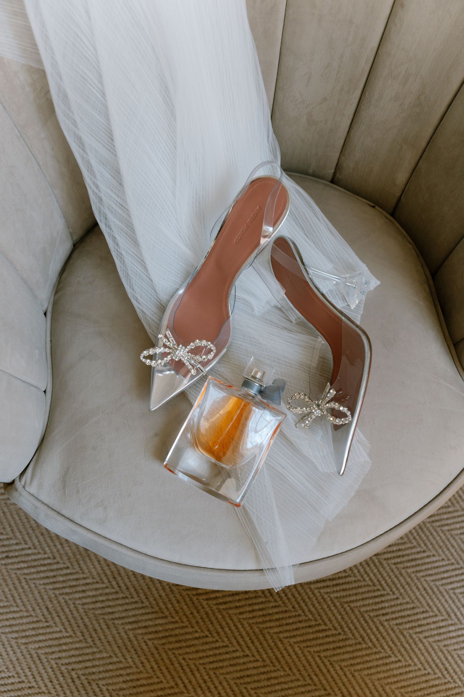 Crystal-adorned clear wedding shoes displayed on a beige chair cushion.