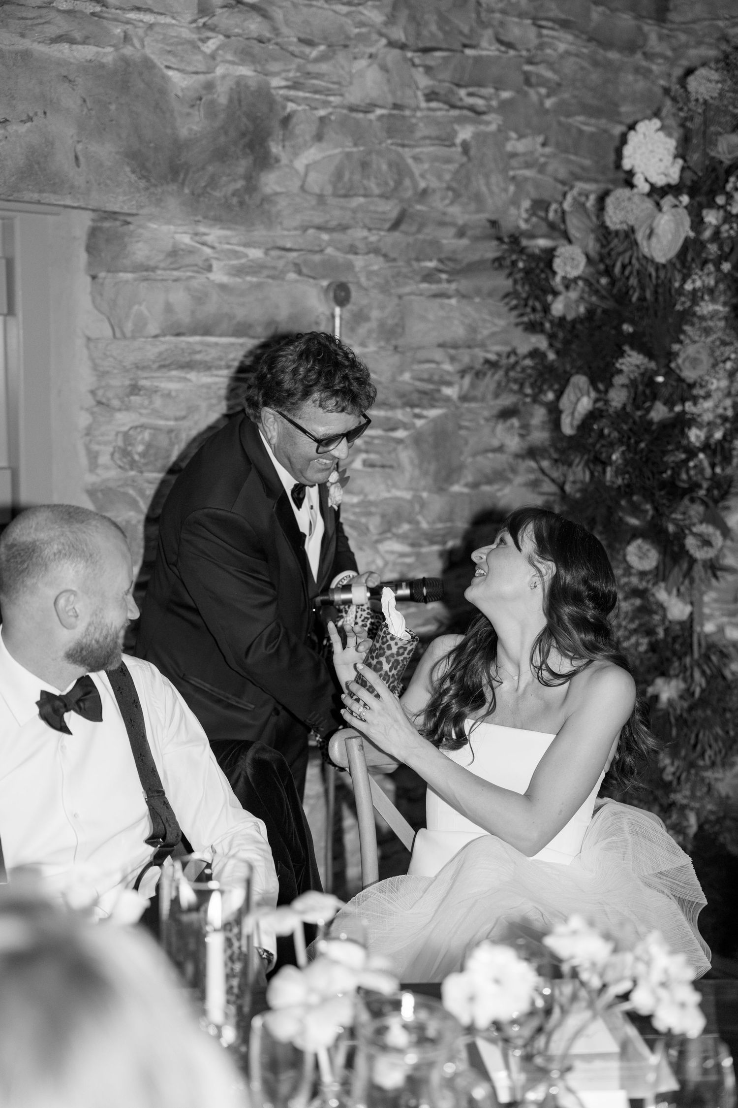 Wedding guests seated at a reception table with stone walls and floral decorations.