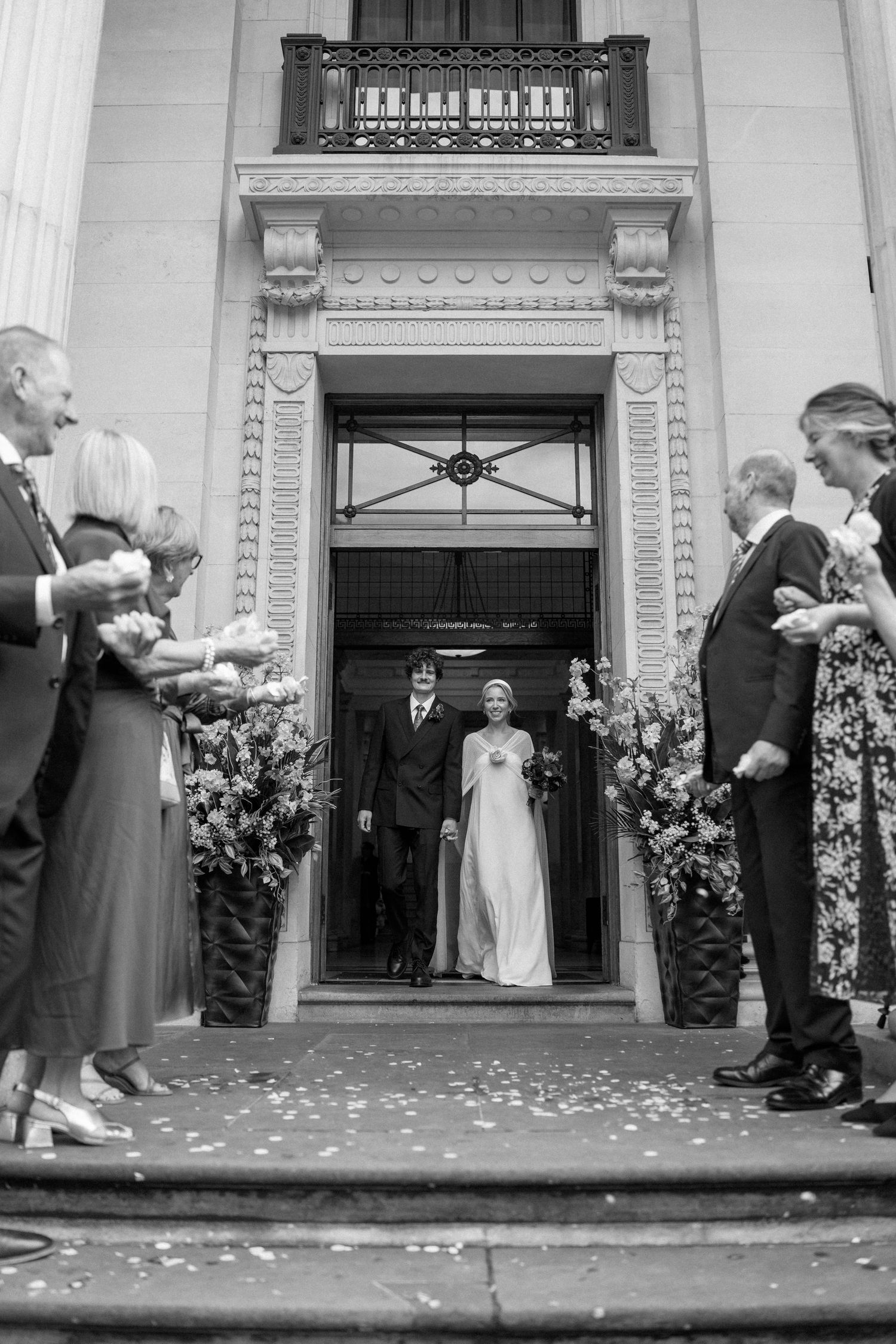 Wedding ceremony exit with guests throwing petals as couple walks through ornate doorway.