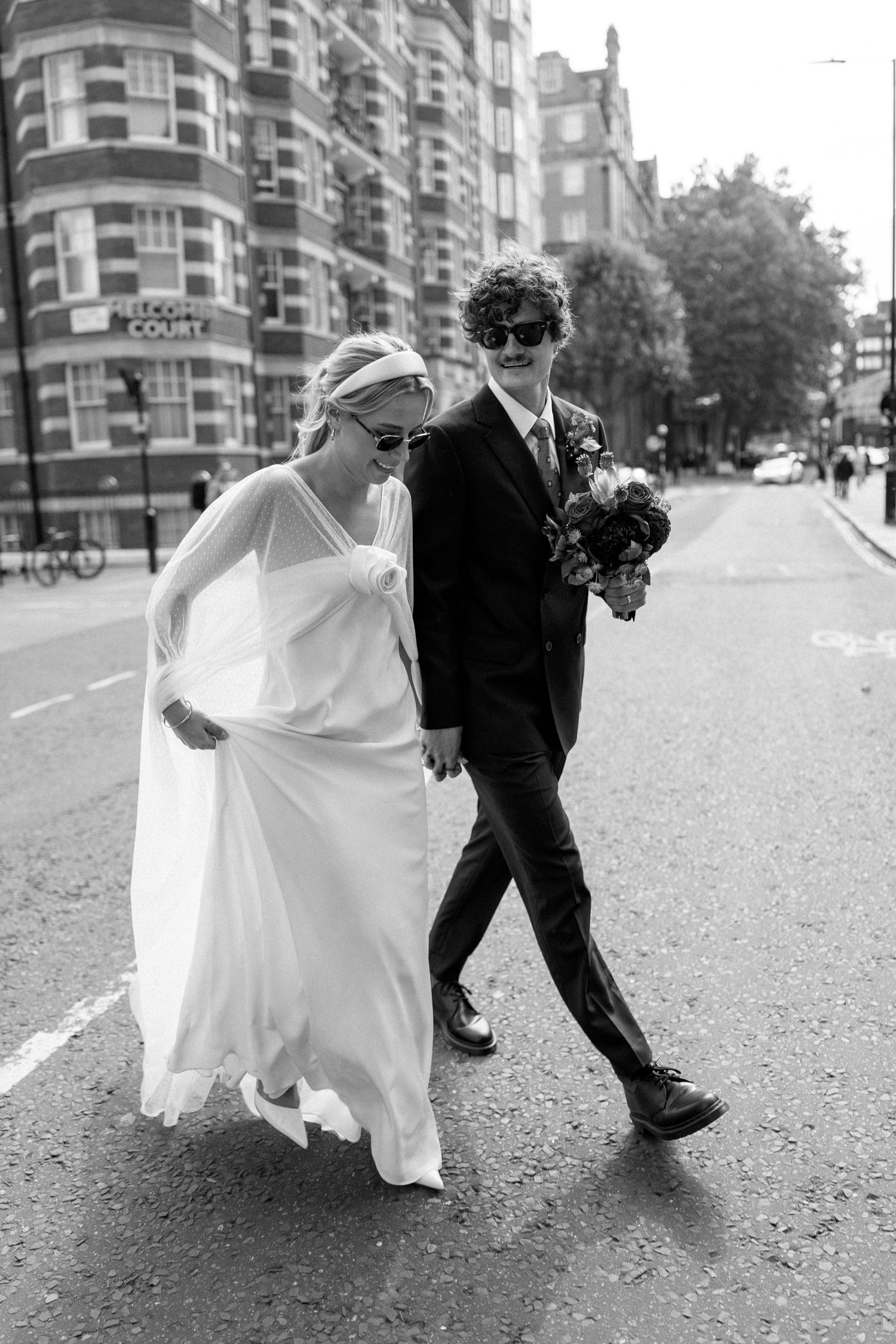 A couple in formal attire walks along a London street with historic buildings in the background.