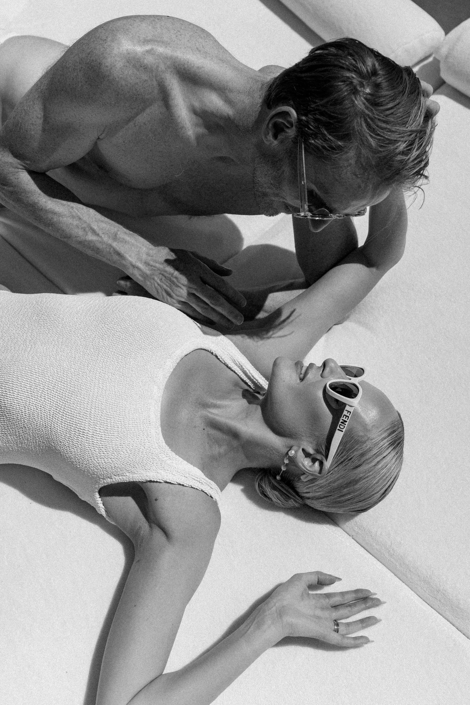 Black and white photo of a couple sunbathing poolside, woman in white swimsuit and sunglasses lying back.