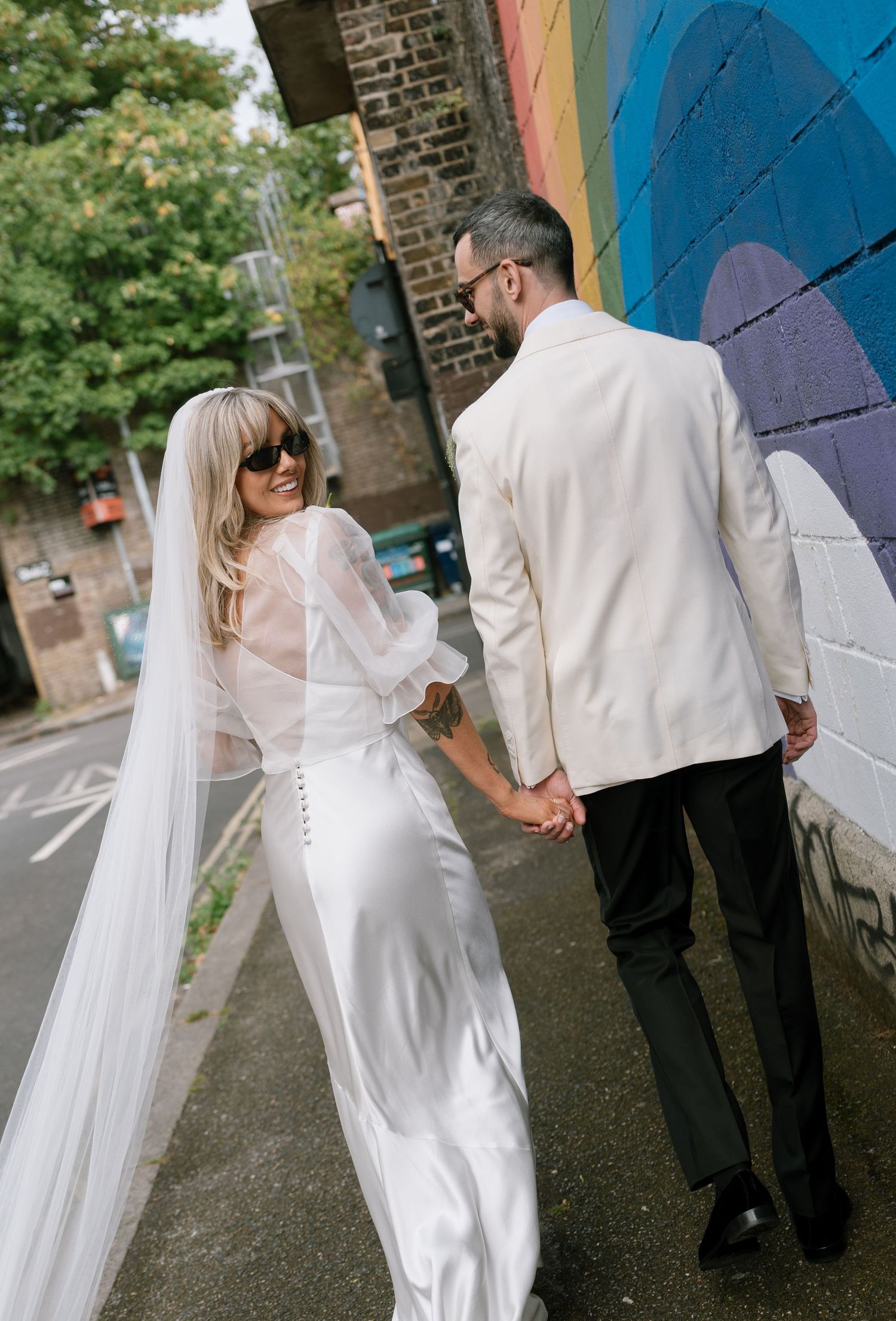 Wedding couple dances and twirls in front of a colorful rainbow mural on a brick wall.