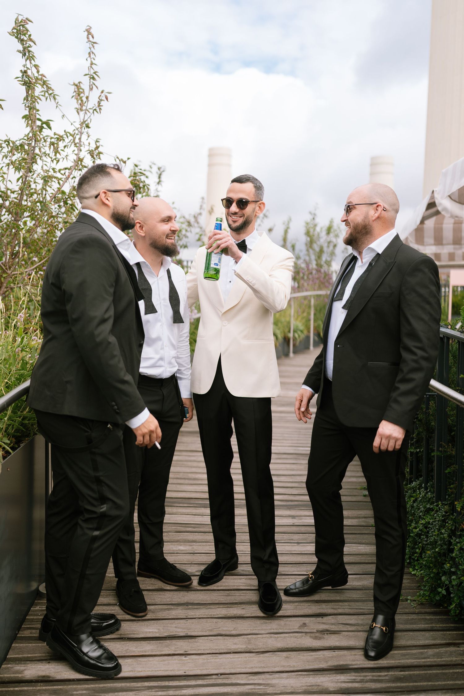 Groomsmen in black suits and sunglasses having fun on a wooden deck outdoors.
