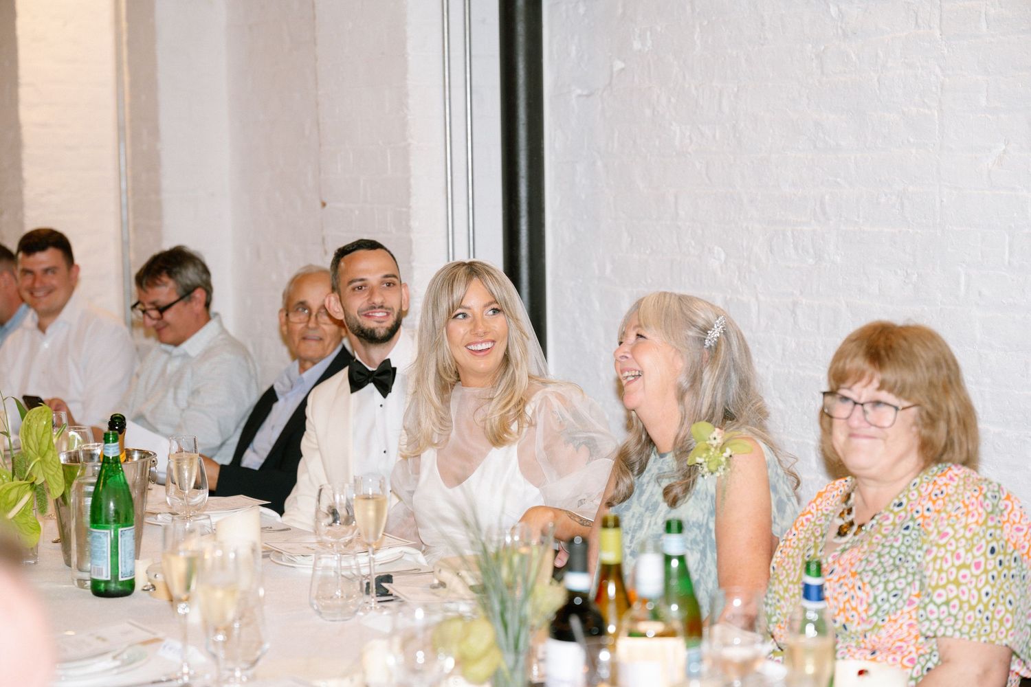 Wedding reception dinner scene with guests seated at a long table with wine bottles and floral centerpieces.
