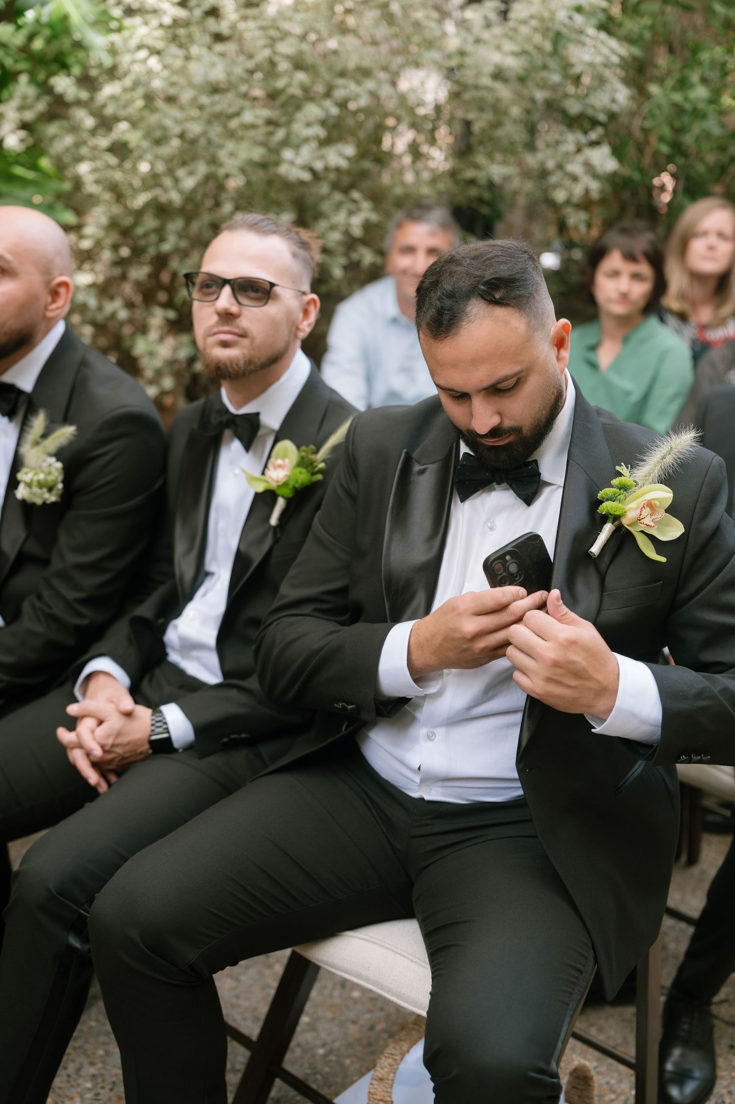 Groomsmen in black tuxedos sit during an outdoor wedding ceremony.