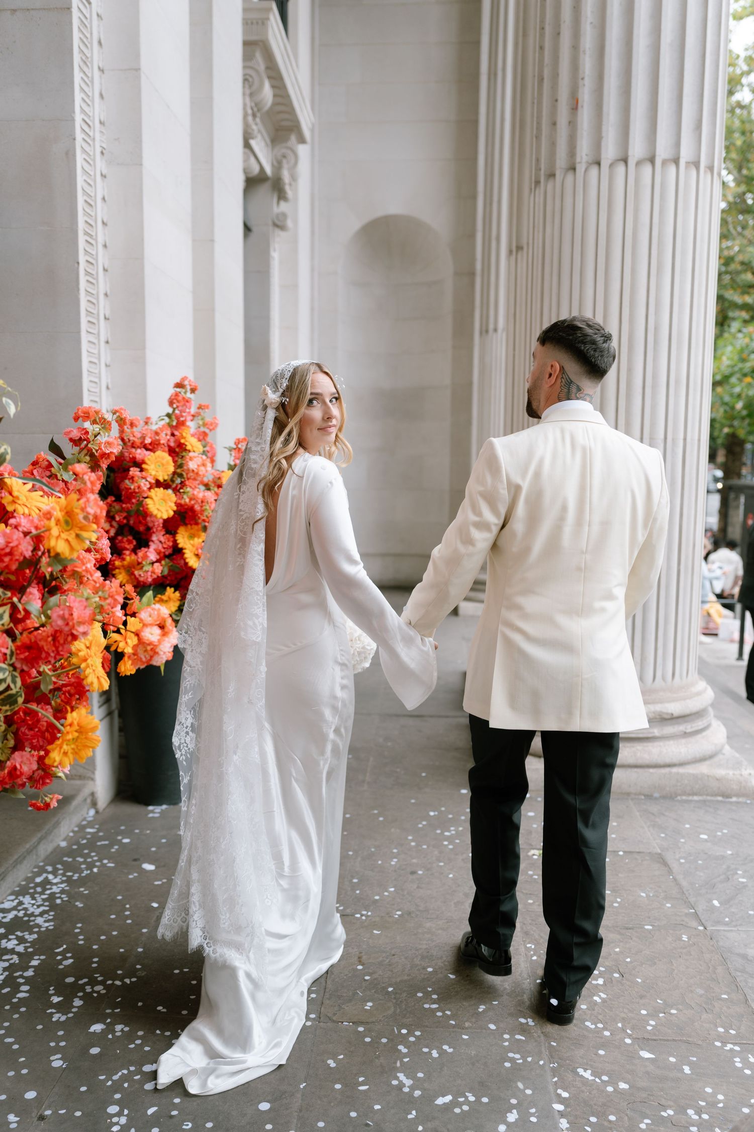 Wedding couple walking hand in hand by classical columns with vibrant orange floral arrangement.