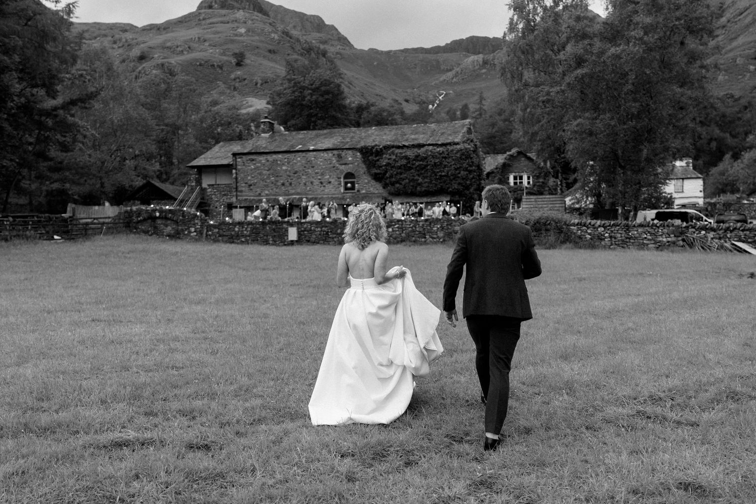 Black and white photo of a couple walking together across a grassy field during a wedding celebration.